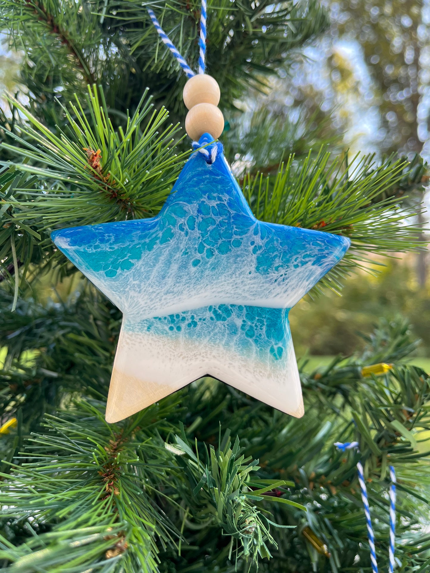 Star-shaped wooden Christmas ornament covered in blue and teal ocean resin art resembling the waves of the ocean. Finished with blue and white baker's twine and two wooden beads