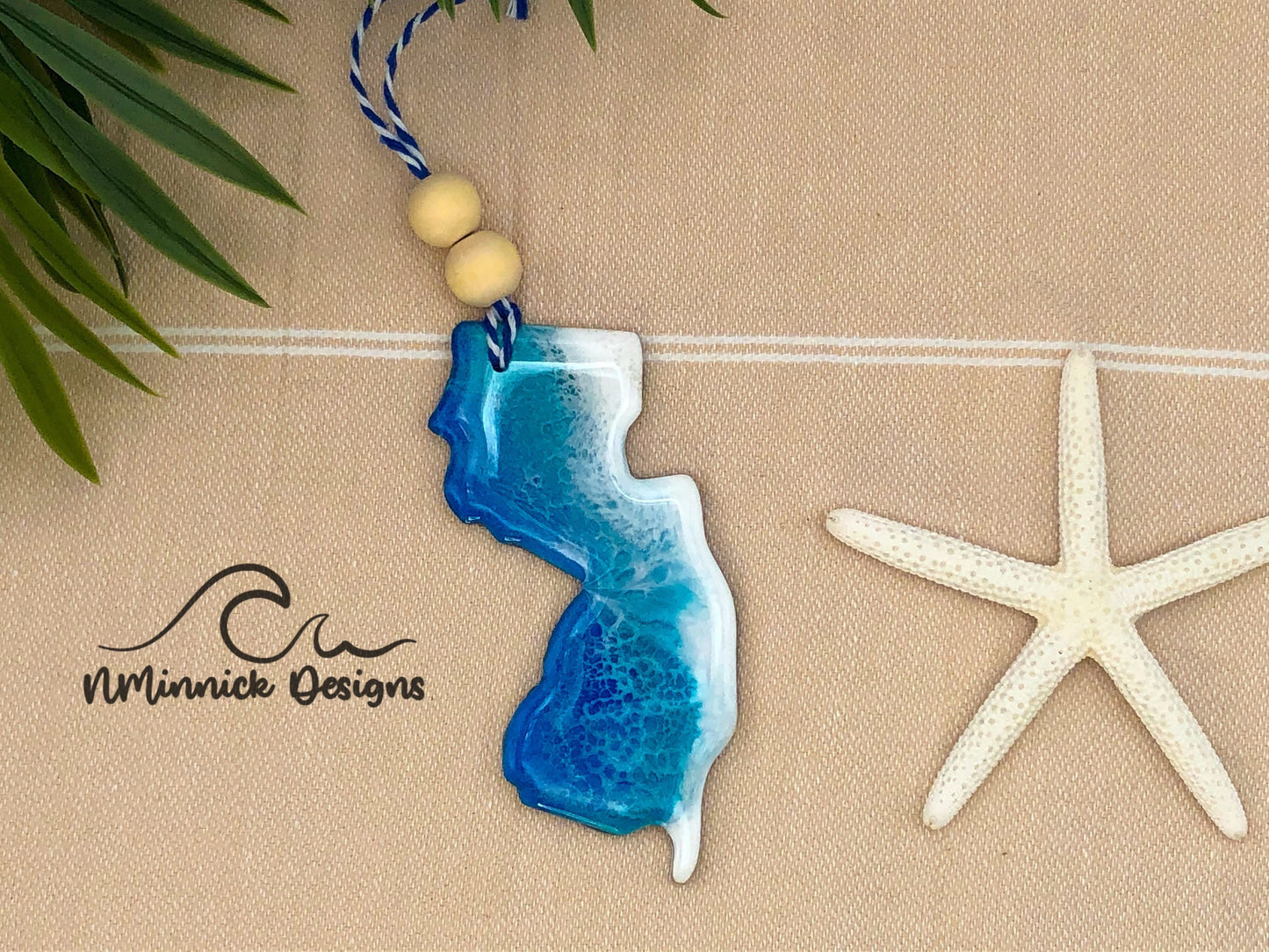 New Jersey-shaped wooden Christmas ornament covered in blue and teal colored ocean resin art resembling the waves along New Jersey's Atlantic Coast. Finished with blue and white baker's twine and two wooden beads.