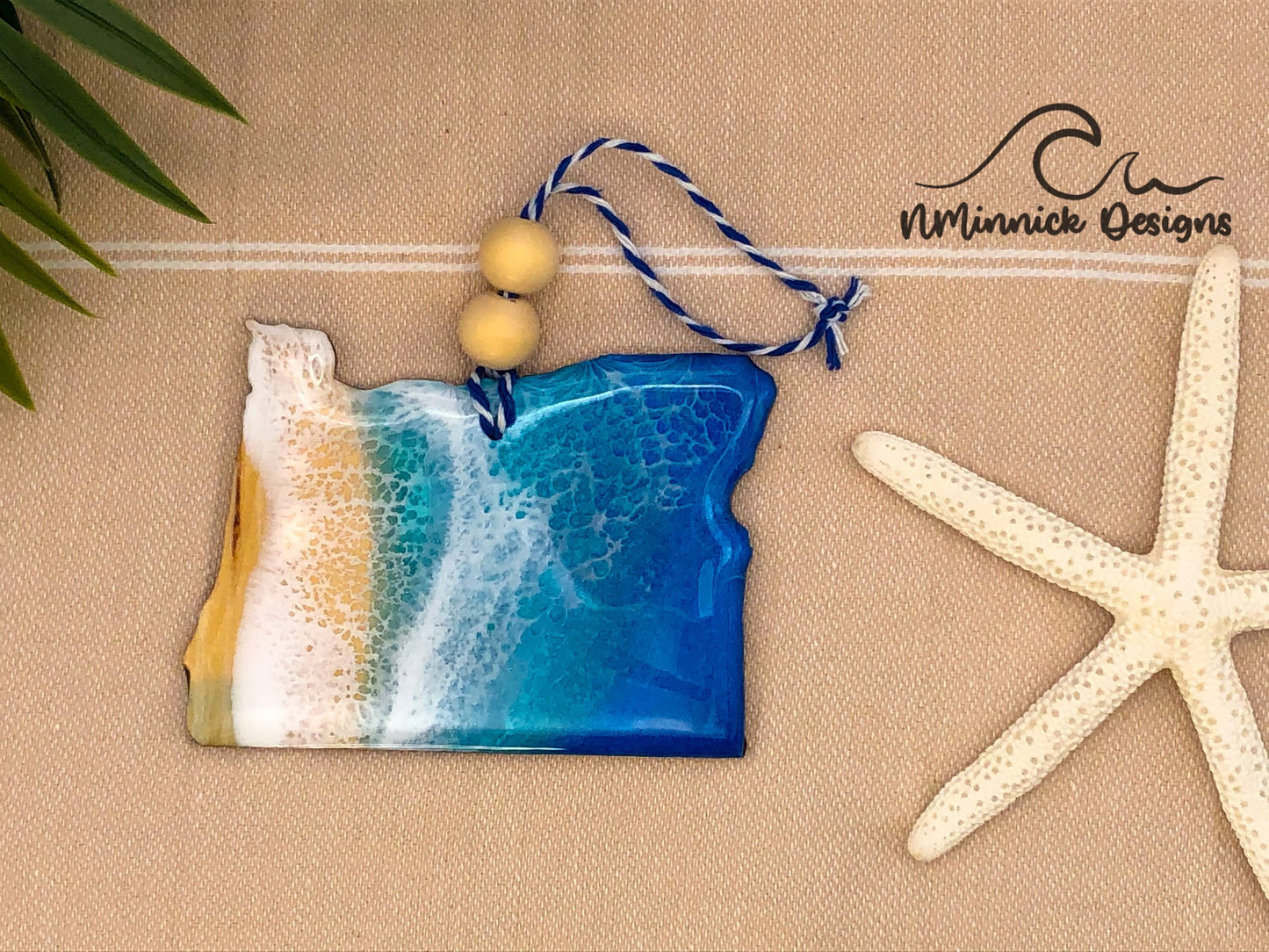 Oregon-shaped wooden Christmas ornament covered in blue and teal colored ocean resin art resembling the waves along Oregon's Pacific Coast. Finished with blue and white baker's twine and two wooden beads.