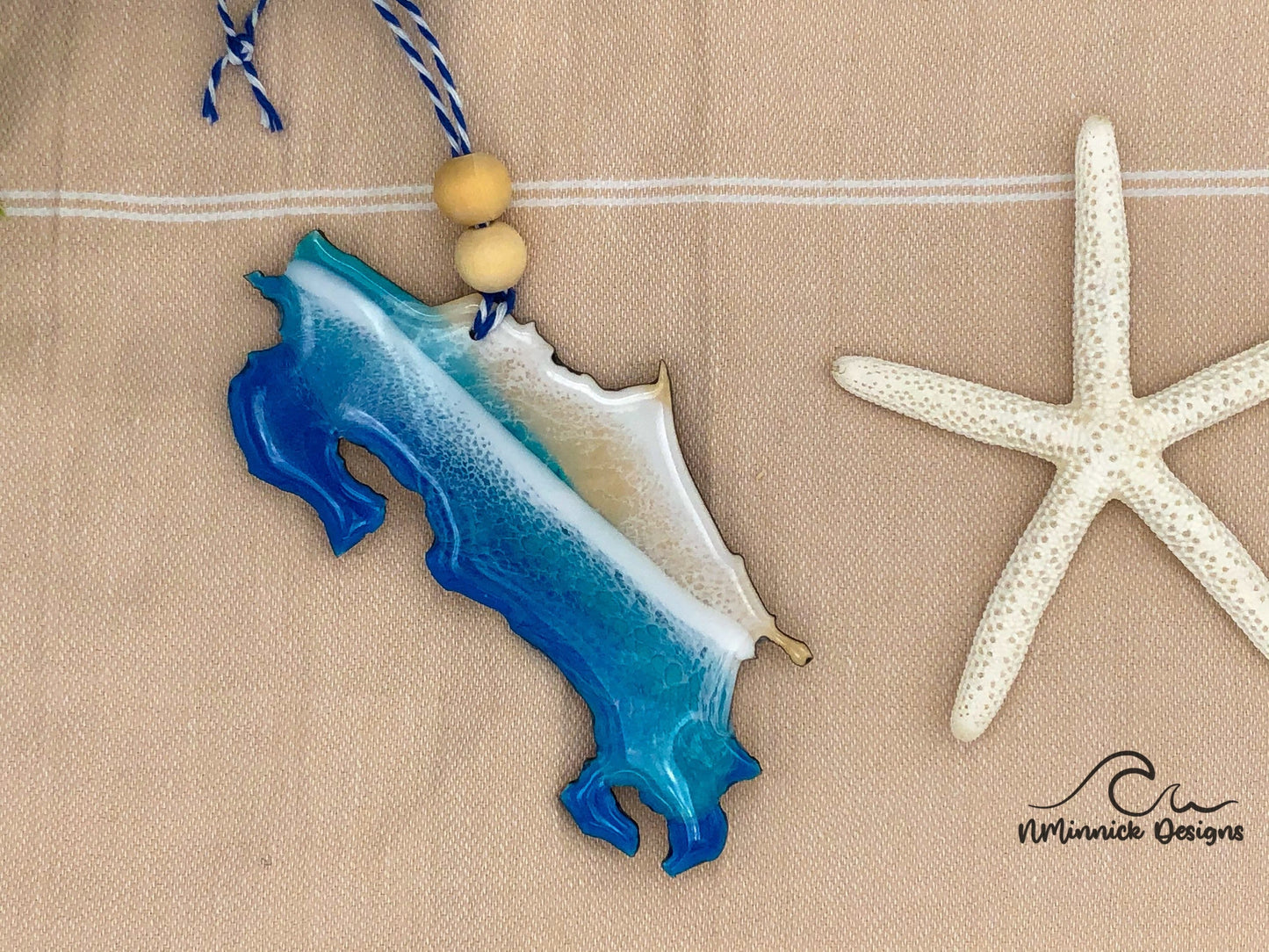 Costa Rica-shaped ornament covered in blue and teal ocean resin art resembling the waves of the ocean. Finished with blue and white baker's twice and two wooden beads.