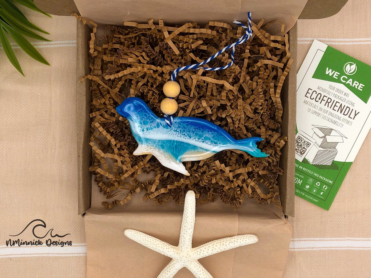 Seal-shaped wooden Christmas ornament covered in blue and teal ocean resin art resembling the waves of the ocean. Finished with blue and white baker's twine and two wooden beads. Laying in kraft colored eco friendly and plastic free packaging.