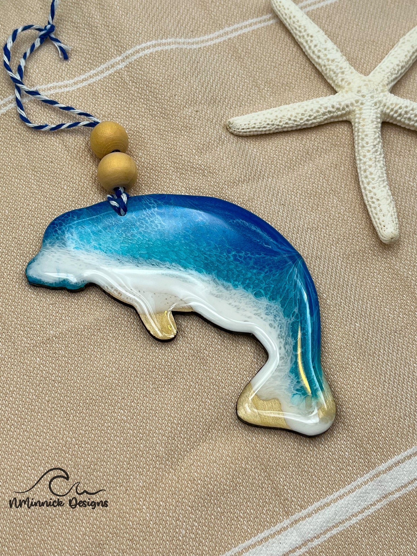 Manatee-shaped wooden Christmas ornament covered in blue and teal ocean resin art resembling the waves of the ocean. Finished with blue and white baker's twine and two wooden beads.