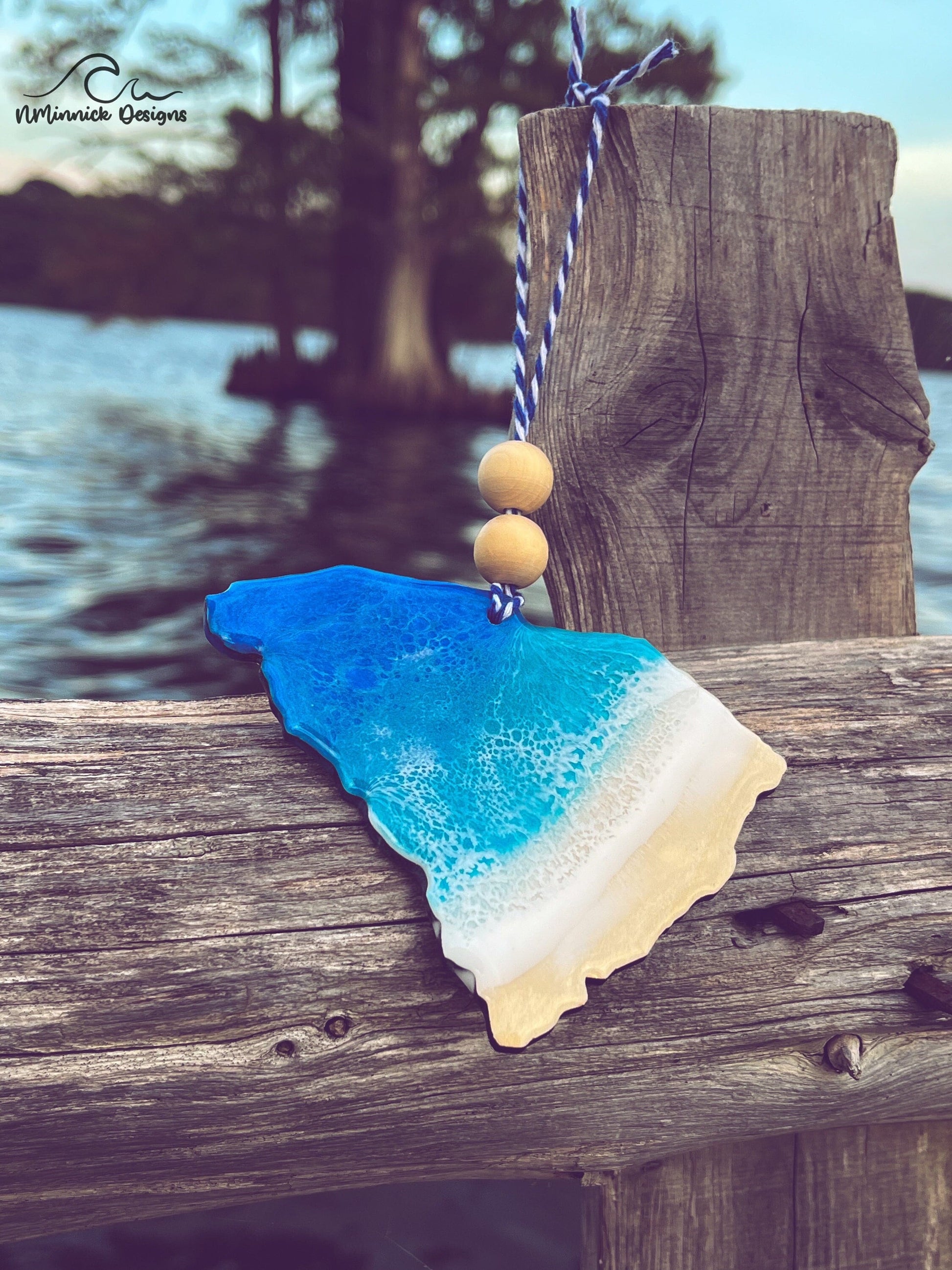 South Carolina-shaped wooden Christmas ornament covered in blue and teal colored ocean resin art resembling the waves along South Carolina's Atlantic Coast. Finished with blue and white baker's twine and two wooden beads.