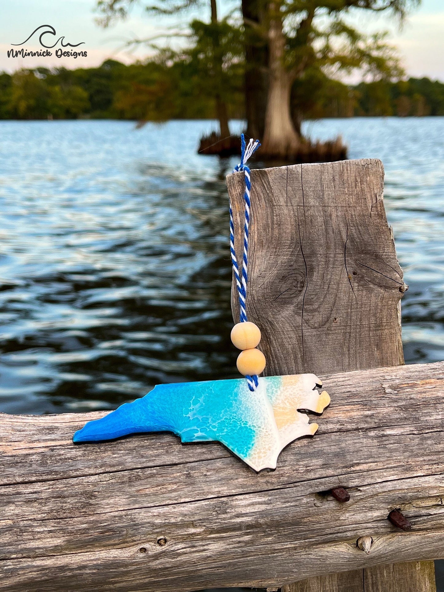 North Carolina-shaped wooden Christmas ornament covered in blue and teal colored ocean resin art resembling the waves along North Carolina's Atlantic Coast. Finished with blue and white baker's twine and two wooden beads.