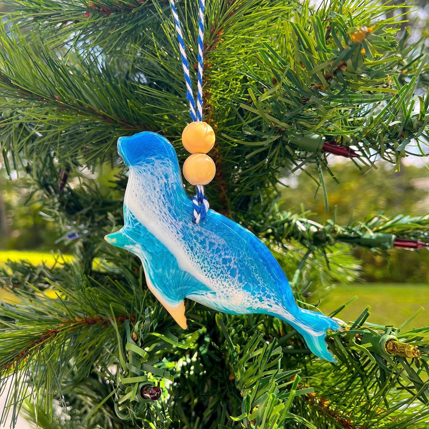 A seal shaped wooden Christmas ornament covered in blue ocean resin art, resembling the waves of the ocean. Finished with baker's twine and two wooden beads and hanging on a Christmas tree.