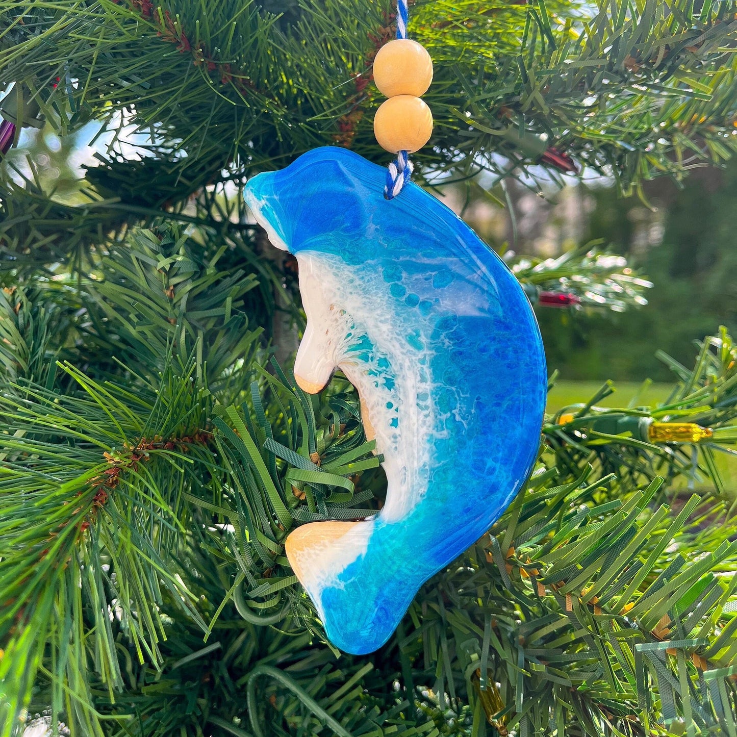 A manatee shaped wooden Christmas ornament covered in blue ocean resin art, resembling the waves of the ocean. Finished with baker's twine and two wooden beads and hanging on a Christmas tree.