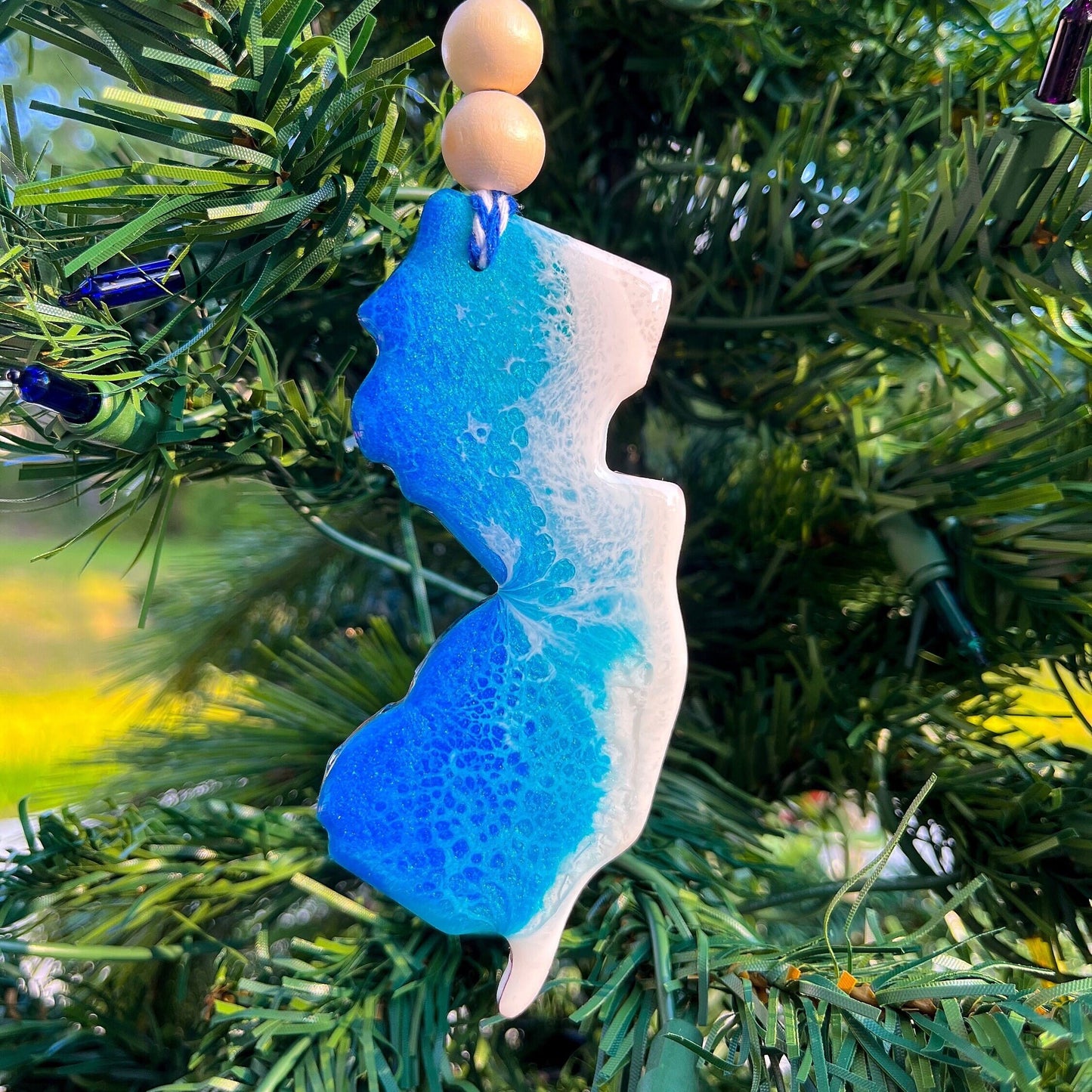New Jersey-shaped wooden Christmas ornament covered in blue and teal colored ocean resin art resembling the waves along New Jersey's Atlantic Coast. Finished with blue and white baker's twine and two wooden beads.