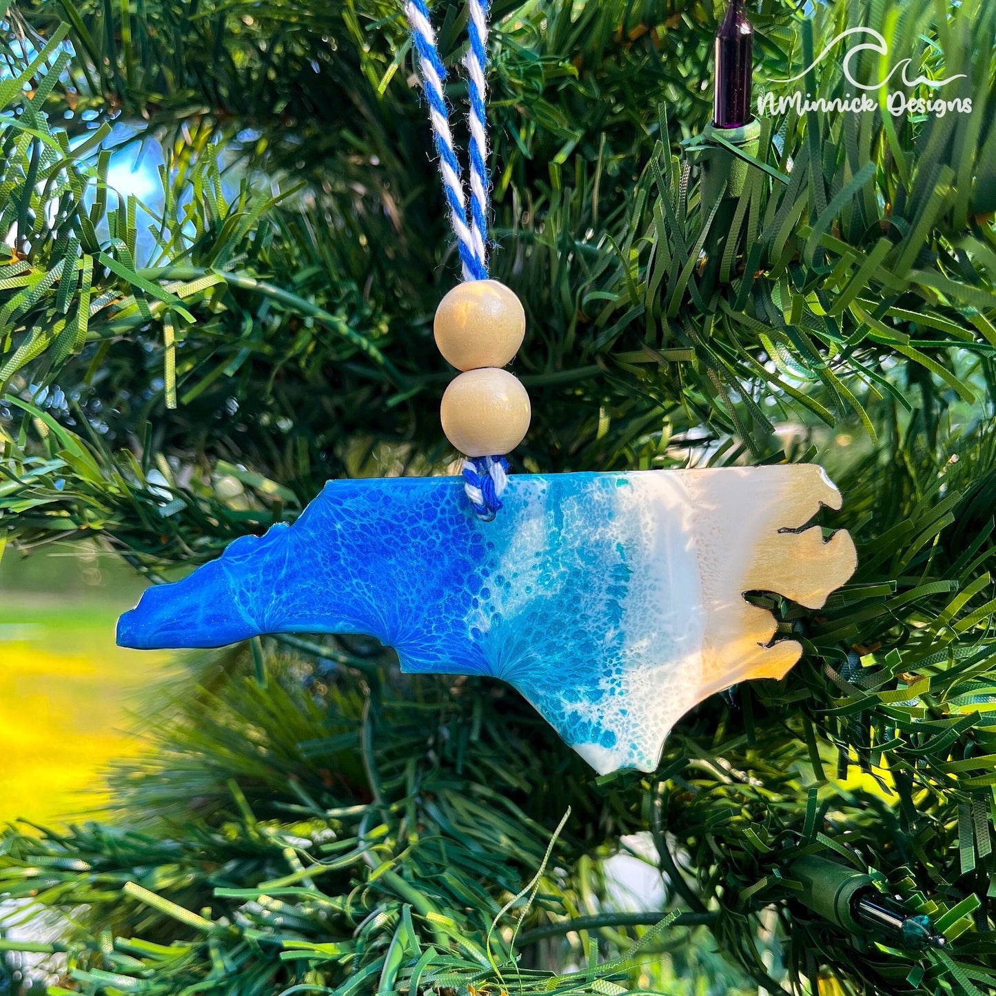 North Carolina-shaped wooden Christmas ornament covered in blue and teal colored ocean resin art resembling the waves along North Carolina's Atlantic Coast. Finished with blue and white baker's twine and two wooden beads.