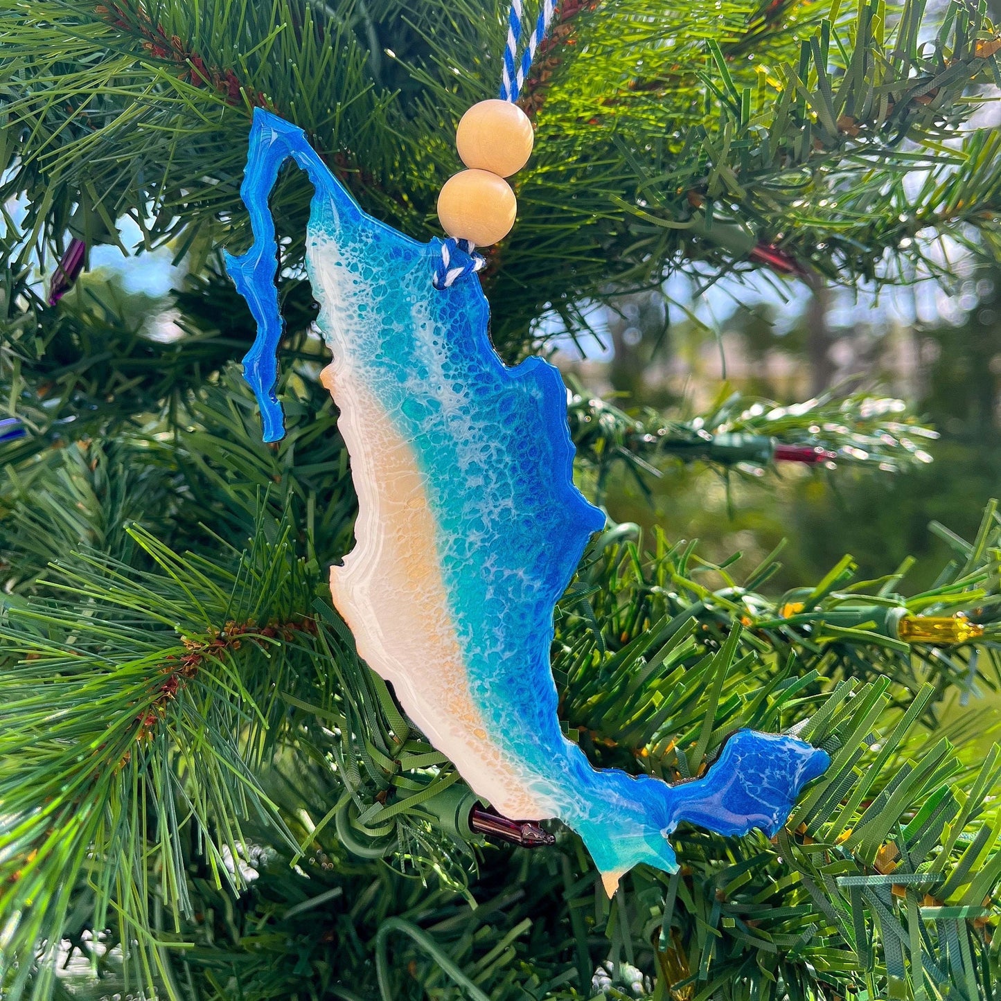 A Mexico shaped wooden Christmas ornament covered in blue ocean resin art, resembling the waves of the ocean.  Finished with baker's twine and two wooden beads and hanging on a Christmas tree.