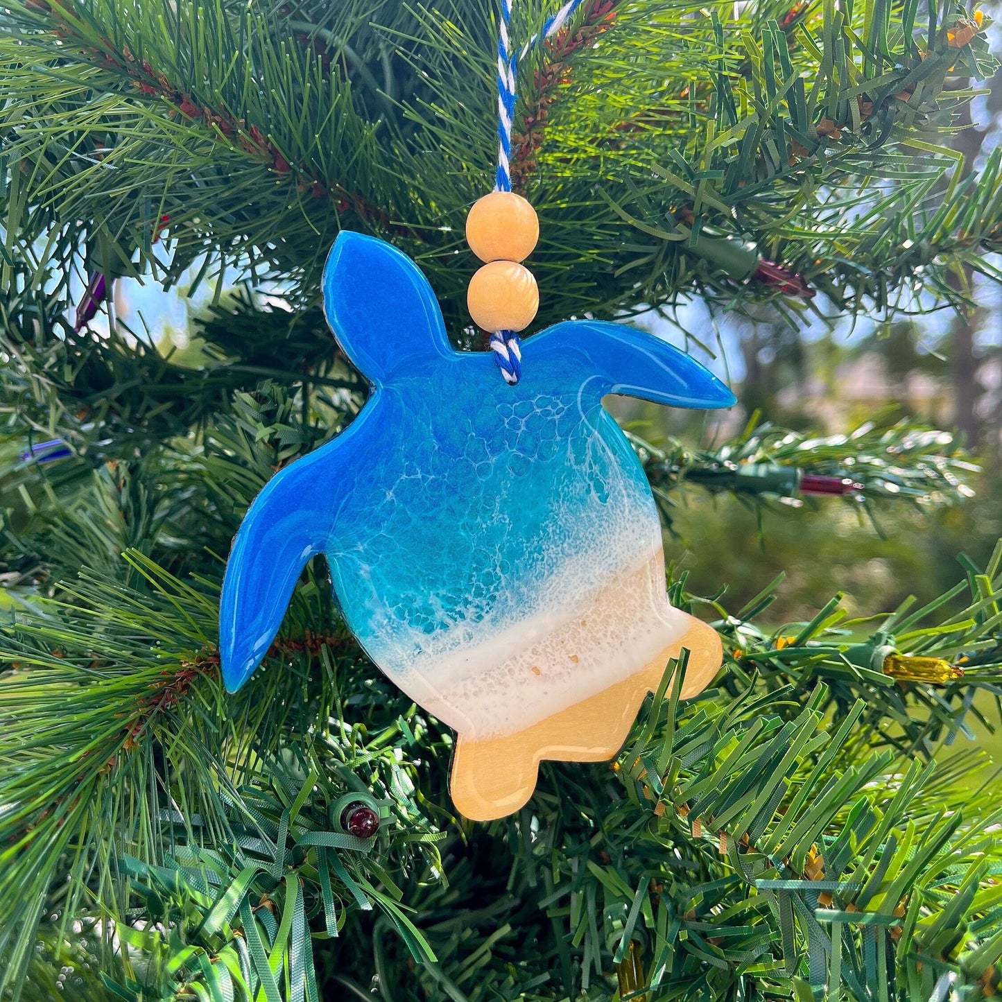 a Sea Turtle shaped wooden Christmas ornament covered in blue ocean resin art, resembling the waves of the ocean. Finished with baker's twine and two wooden beads and hanging on a Christmas tree.