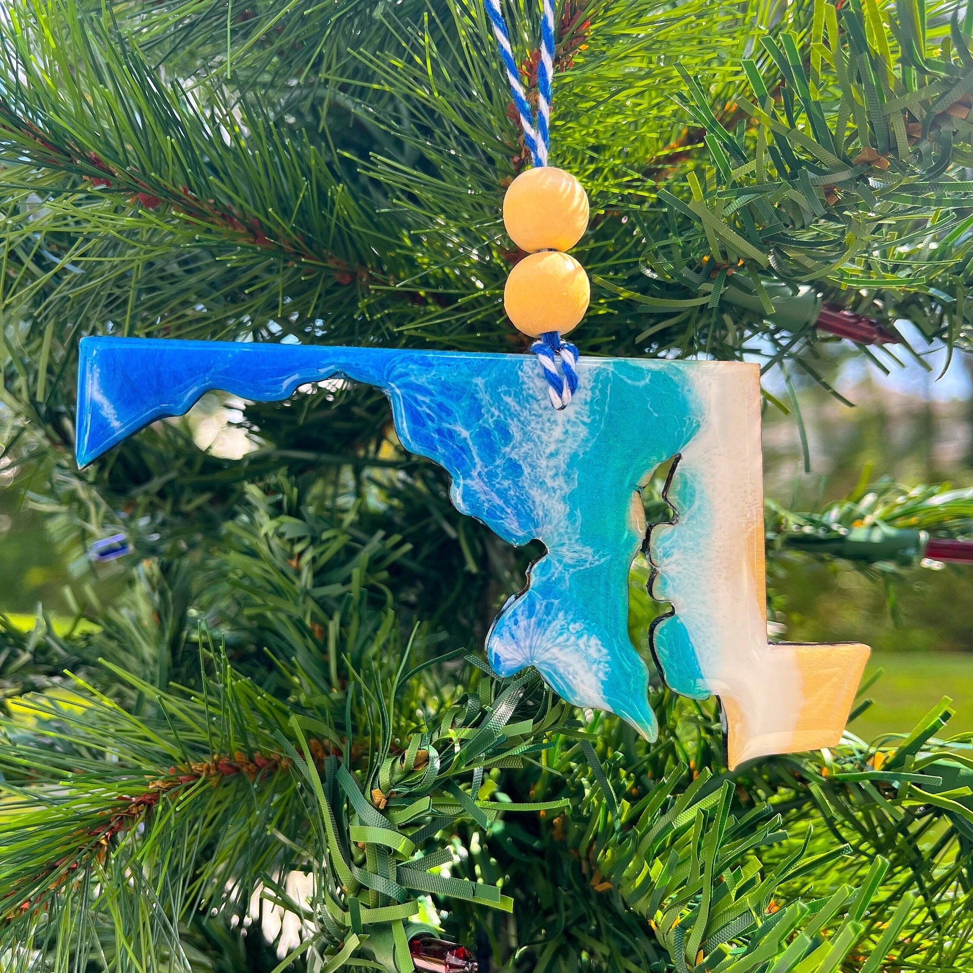 A Maryland shaped wooden Christmas ornament covered in blue ocean resin art, resembling the waves of the ocean.  Finished with baker's twine and two wooden beads and hanging on a Christmas tree.