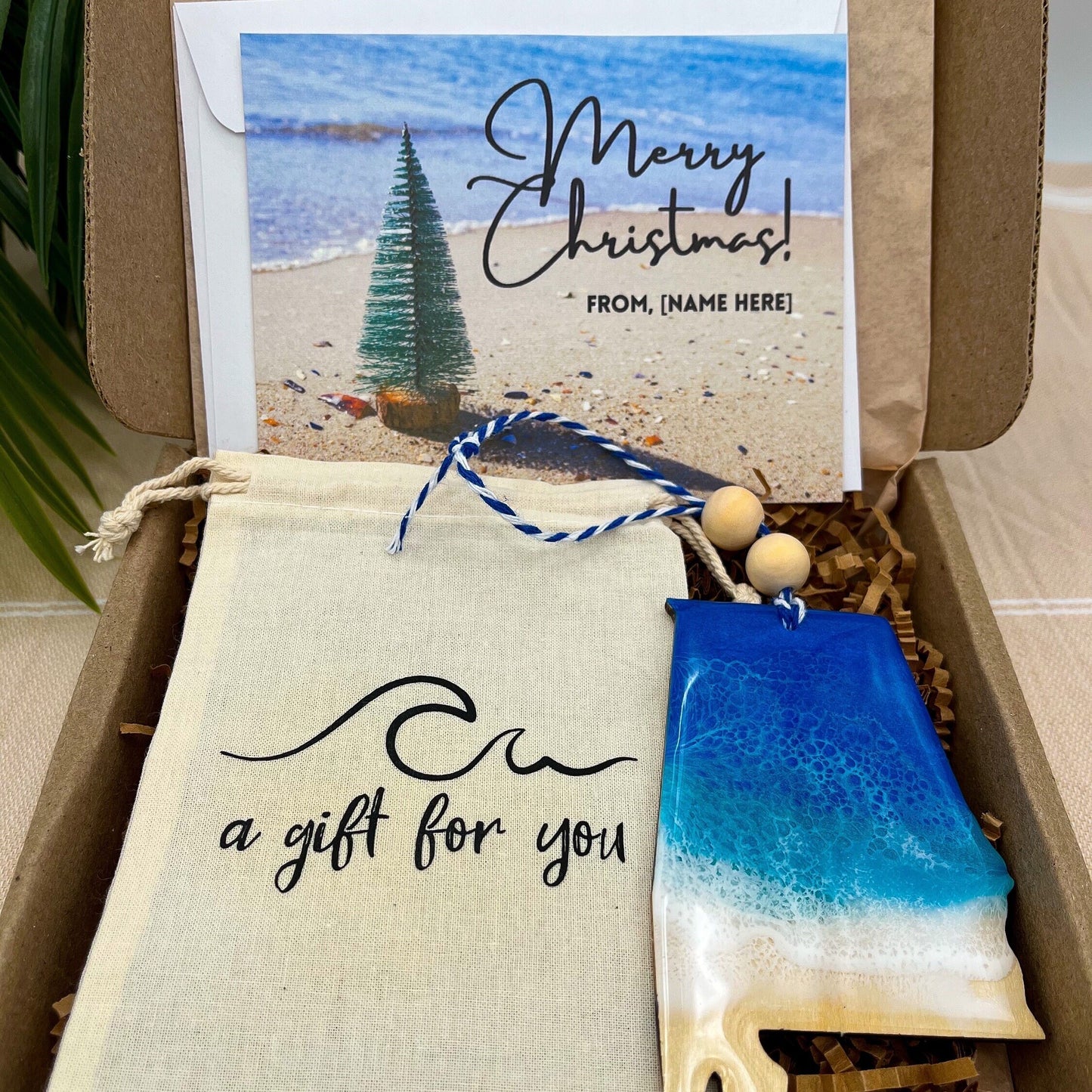 An Alabama shaped wooden christmas ornament with ocean resin art, resembling the waves of the ocean. In a box with an ornament keepsake bag with a wave that says "a gift for you" and a Merry Christmas custom card.