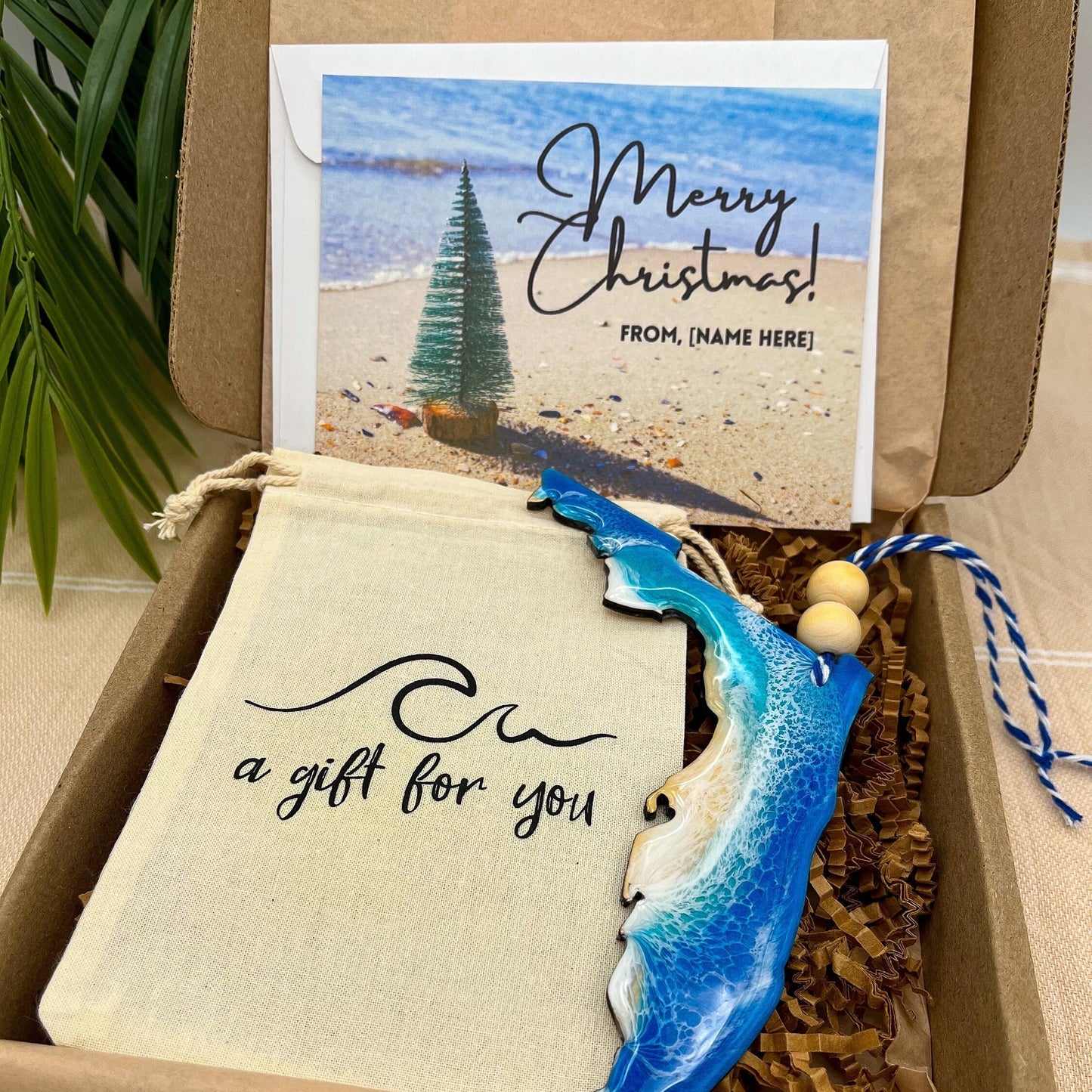 A Florida shaped wooden christmas ornament with ocean resin art, resembling the waves of the ocean. In a box with an ornament keepsake bag with a wave that says "a gift for you" and a Merry Christmas custom card.