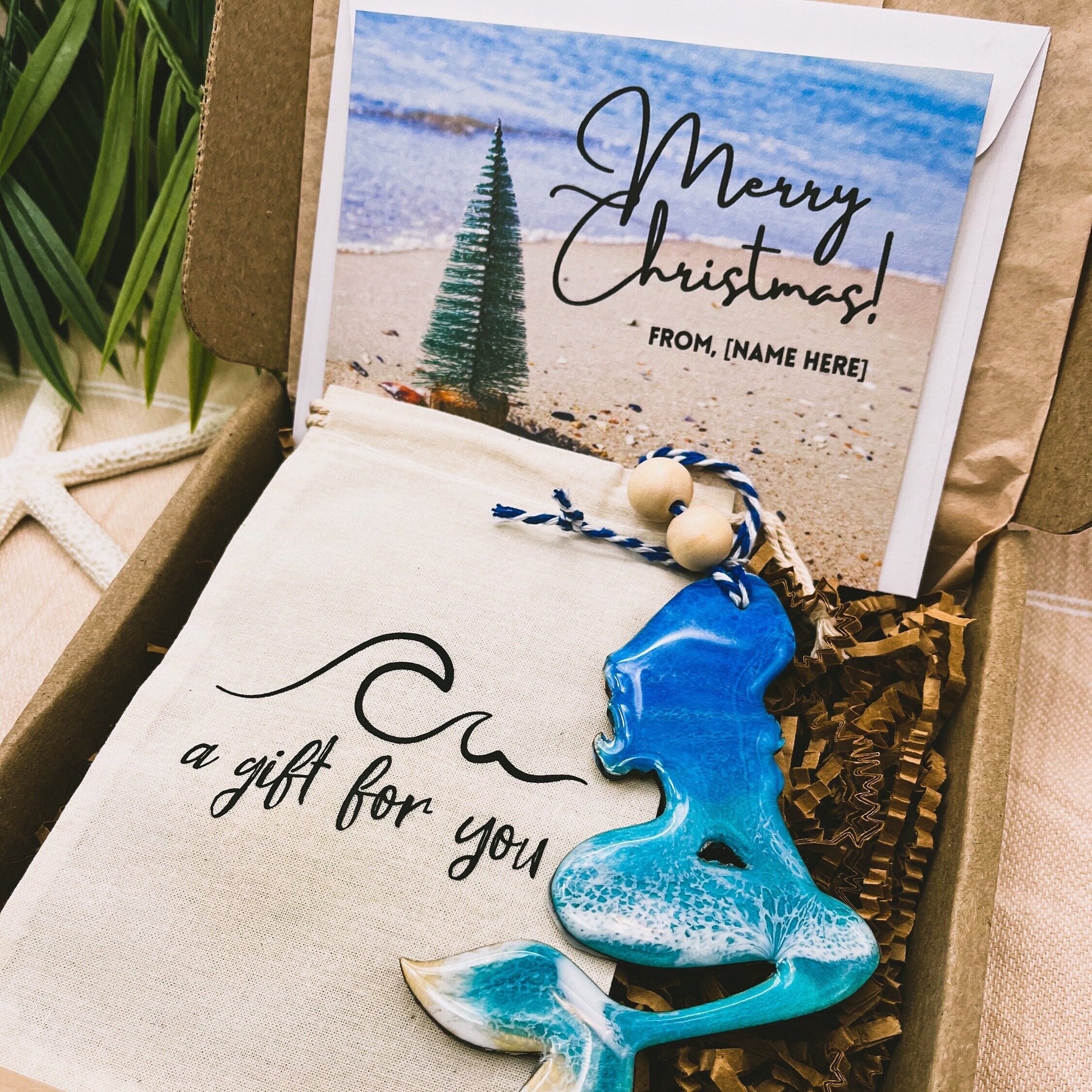 A Mermaid shaped wooden christmas ornament with ocean resin art, resembling the waves of the ocean. In a box with an ornament keepsake bag with a wave that says "a gift for you" and a Merry Christmas custom card.