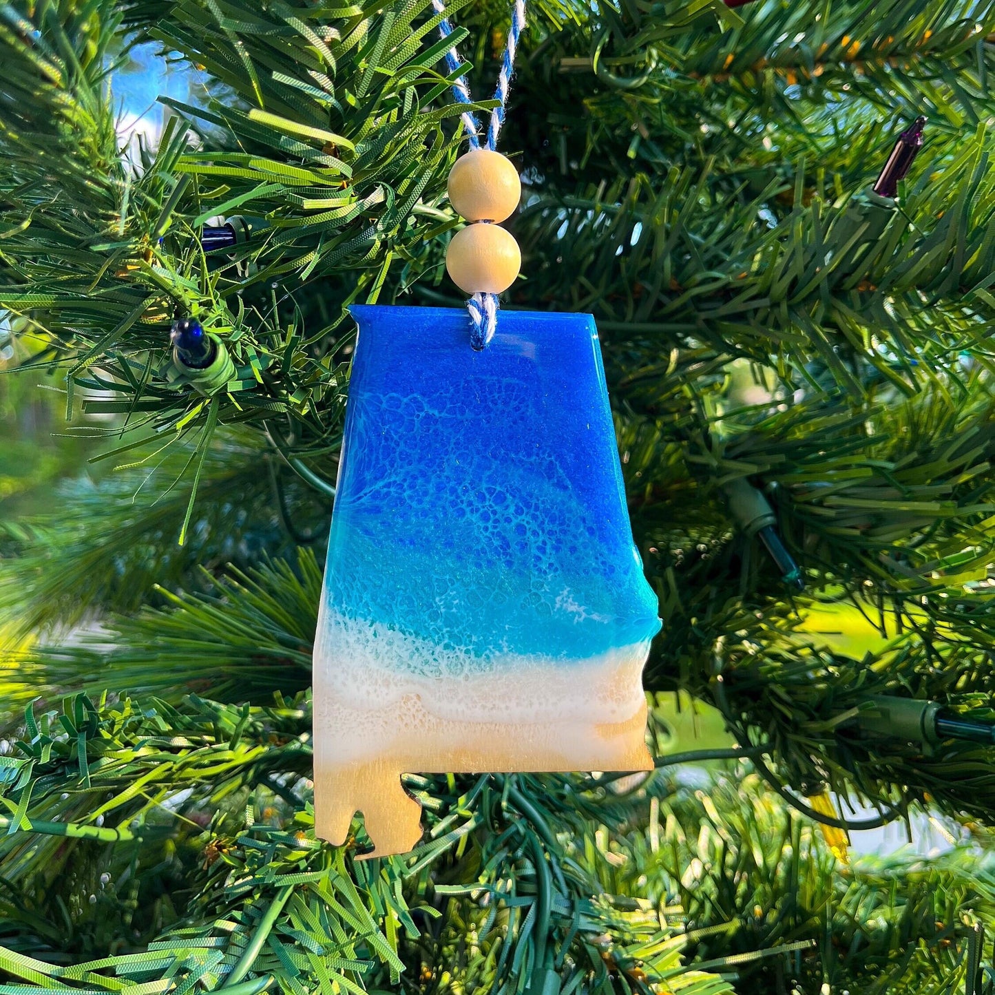 Alabama-shaped wooden Christmas ornament covered in blue and teal colored ocean resin art resembling the waves along Alabama's Gulf Coast. Finished with blue and white baker's twine and two wooden beads.