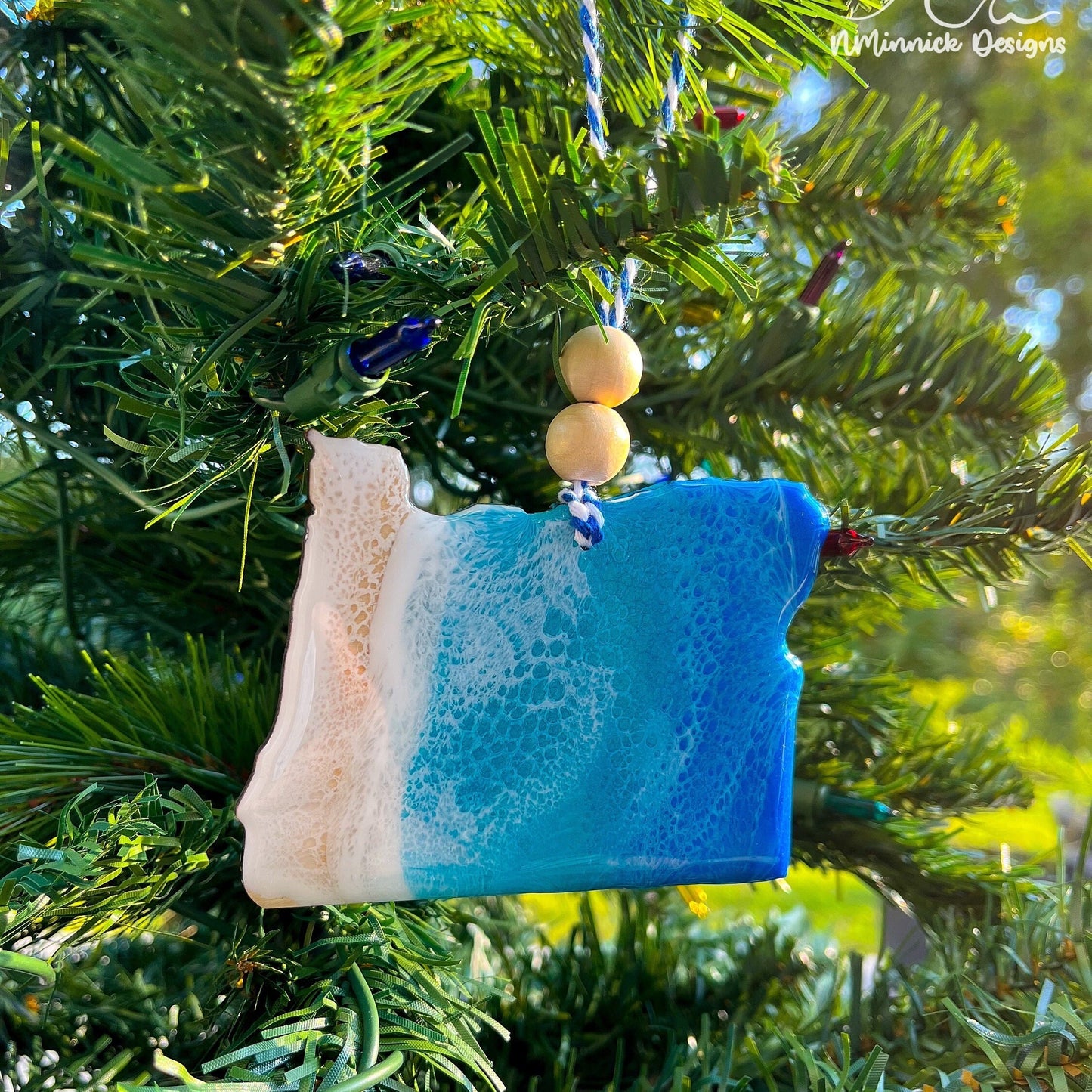 Oregon-shaped wooden Christmas ornament covered in blue and teal colored ocean resin art resembling the waves along Oregon's Pacific Coast. Finished with blue and white baker's twine and two wooden beads.