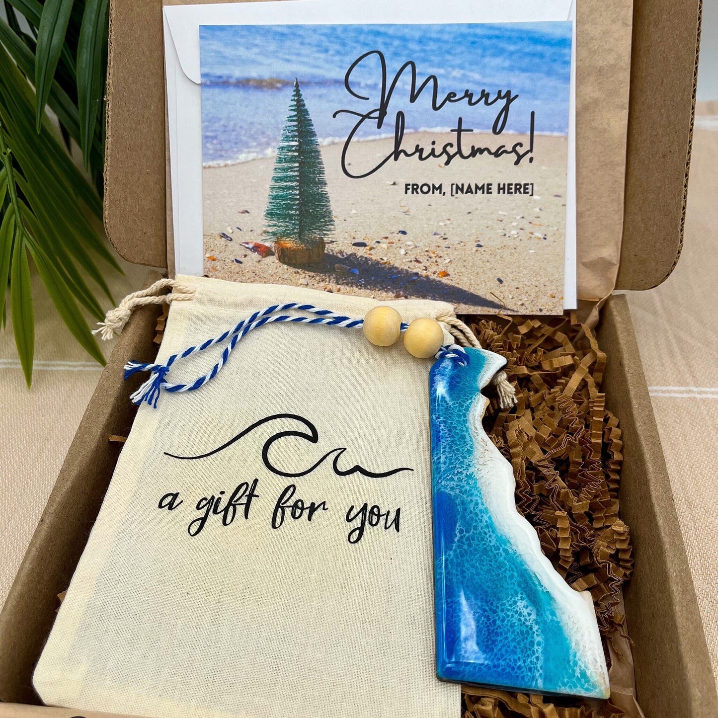 A Delaware shaped wooden christmas ornament with ocean resin art, resembling the waves of the ocean. In a box with an ornament keepsake bag with a wave that says "a gift for you" and a Merry Christmas custom card.
