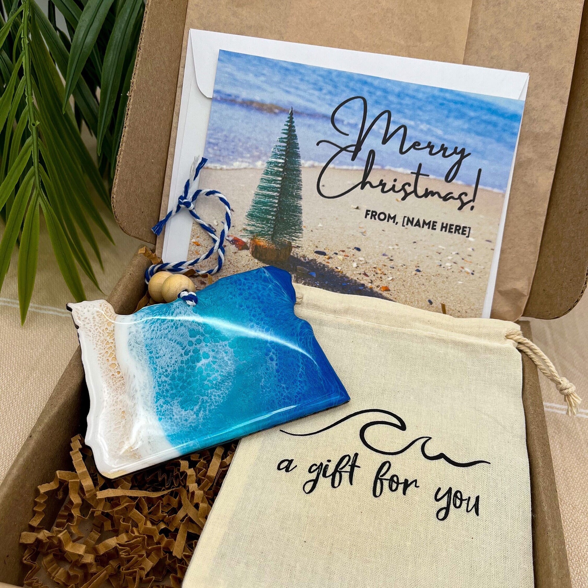 An Oregon shaped wooden christmas ornament with ocean resin art, resembling the waves of the ocean. In a box with an ornament keepsake bag with a wave that says "a gift for you" and a Merry Christmas custom card.