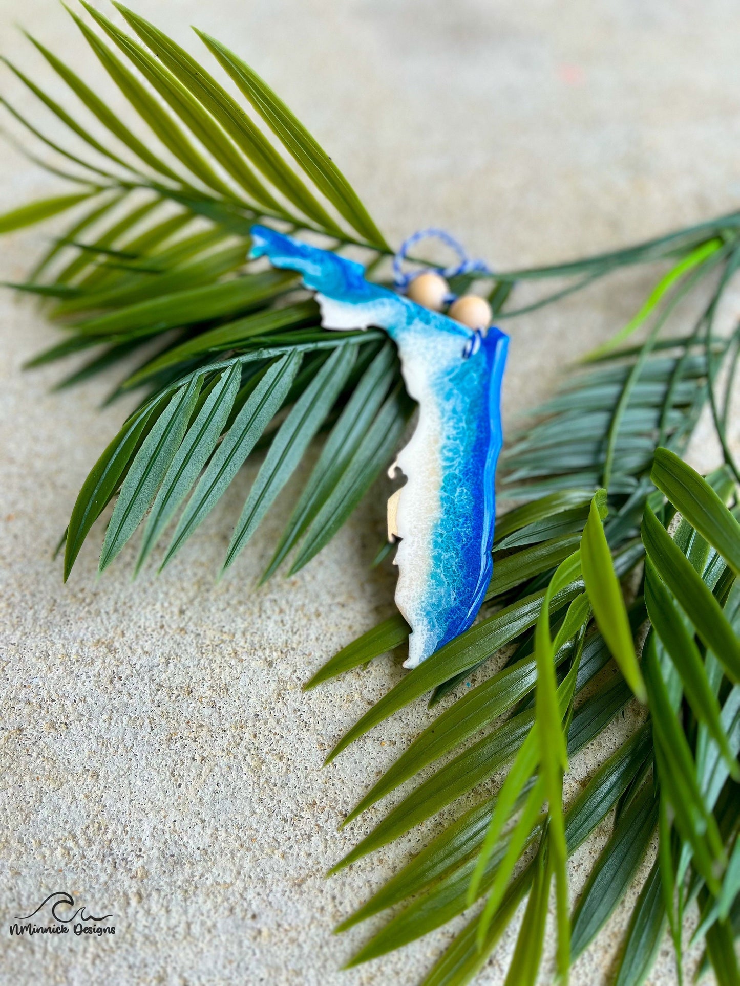 A florida shaped wooden ornament with blue and white ocean resin art. Finished with two wooden beads and a stand of bakers twine.