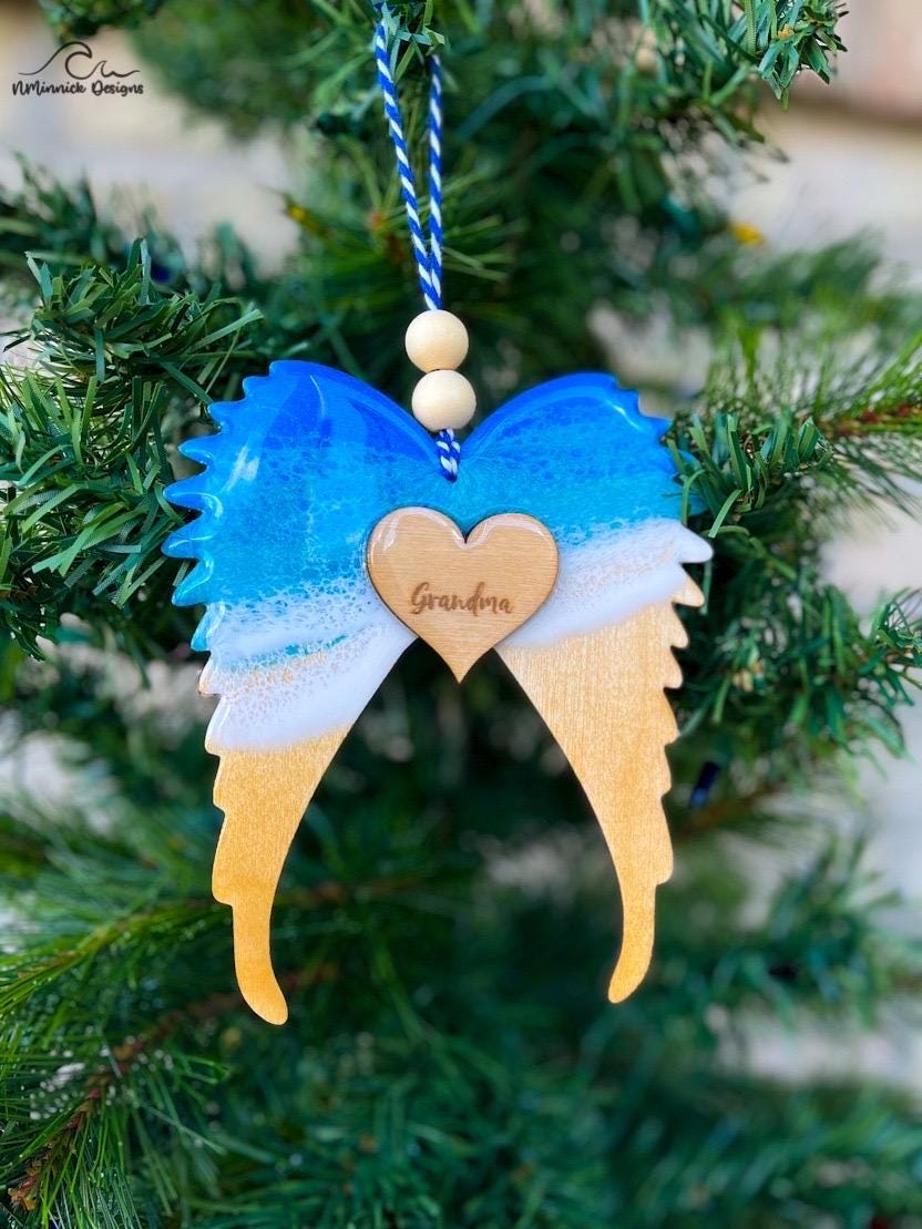 An angel wings memorial keepsake remembrance ornament adorned with ocean resin waves and a heart pendant personalized with the name of a late loved one.