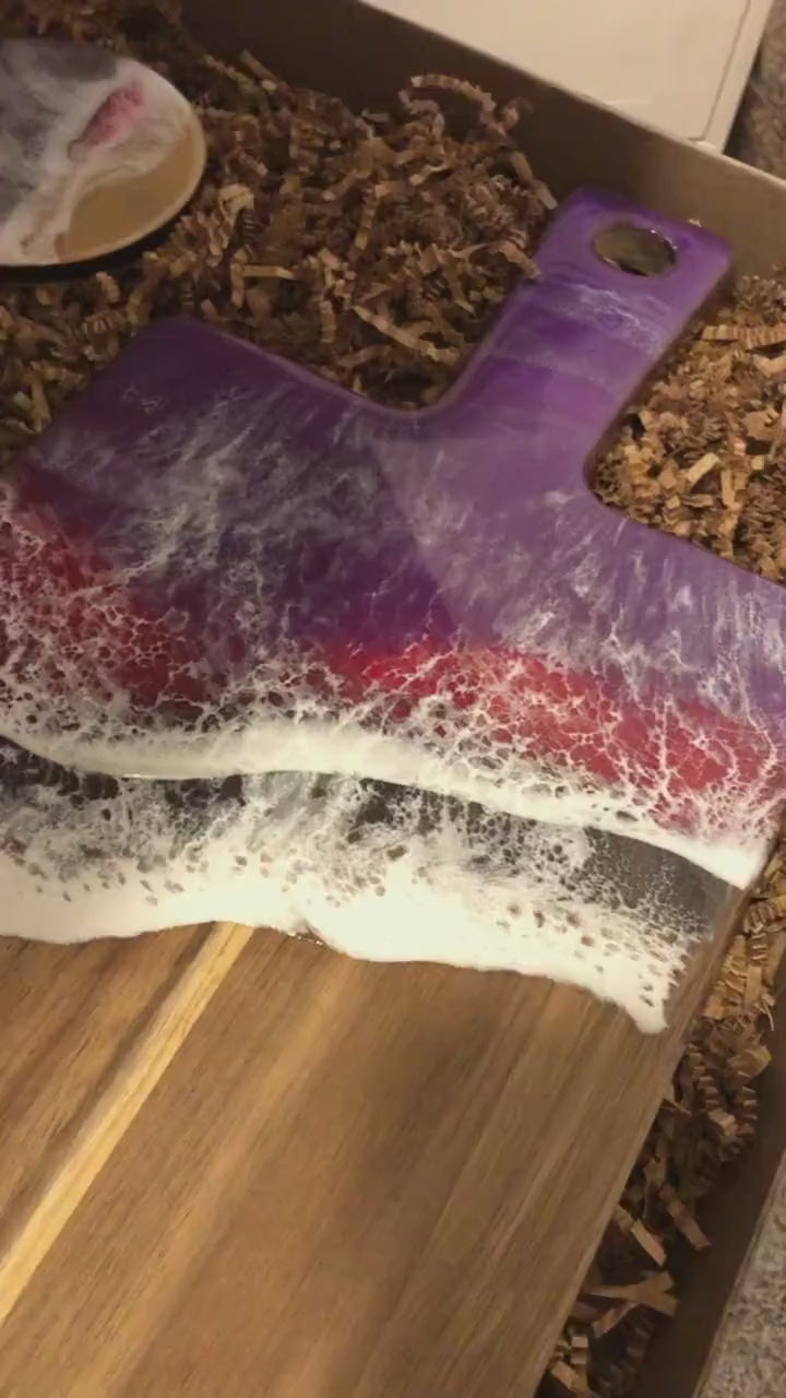 Long Acacia Wood Serving Board with Handle, top half covered in purple, red, and black ocean resin art resembling the waves of the ocean. Matching set of 4 ocean resin art coated drink coasters with cork backings shown laying in a kraft colored eco-friendly and plastic free gift box packaging.