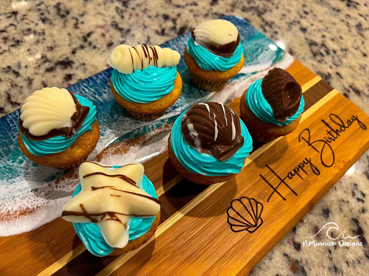 Bamboo Serving Tray laser engraved with a seashell and Happy Birthday on the bottom and covered in blue and teal ocean resin art that resembles the waves of the ocean. Decorated with cupcakes with blue icing and adorned with chocolate seashells.