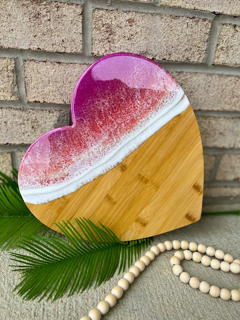 heart shaped charcuterie board with magenta, coral, and golden colored ocean resin waves on top. Setting against a white brick background with palm leaves and wooden beads.