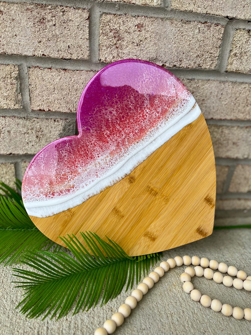 heart shaped charcuterie board with magenta, coral, and golden colored ocean resin waves on top. Setting against a white brick background with palm leaves and wooden beads.