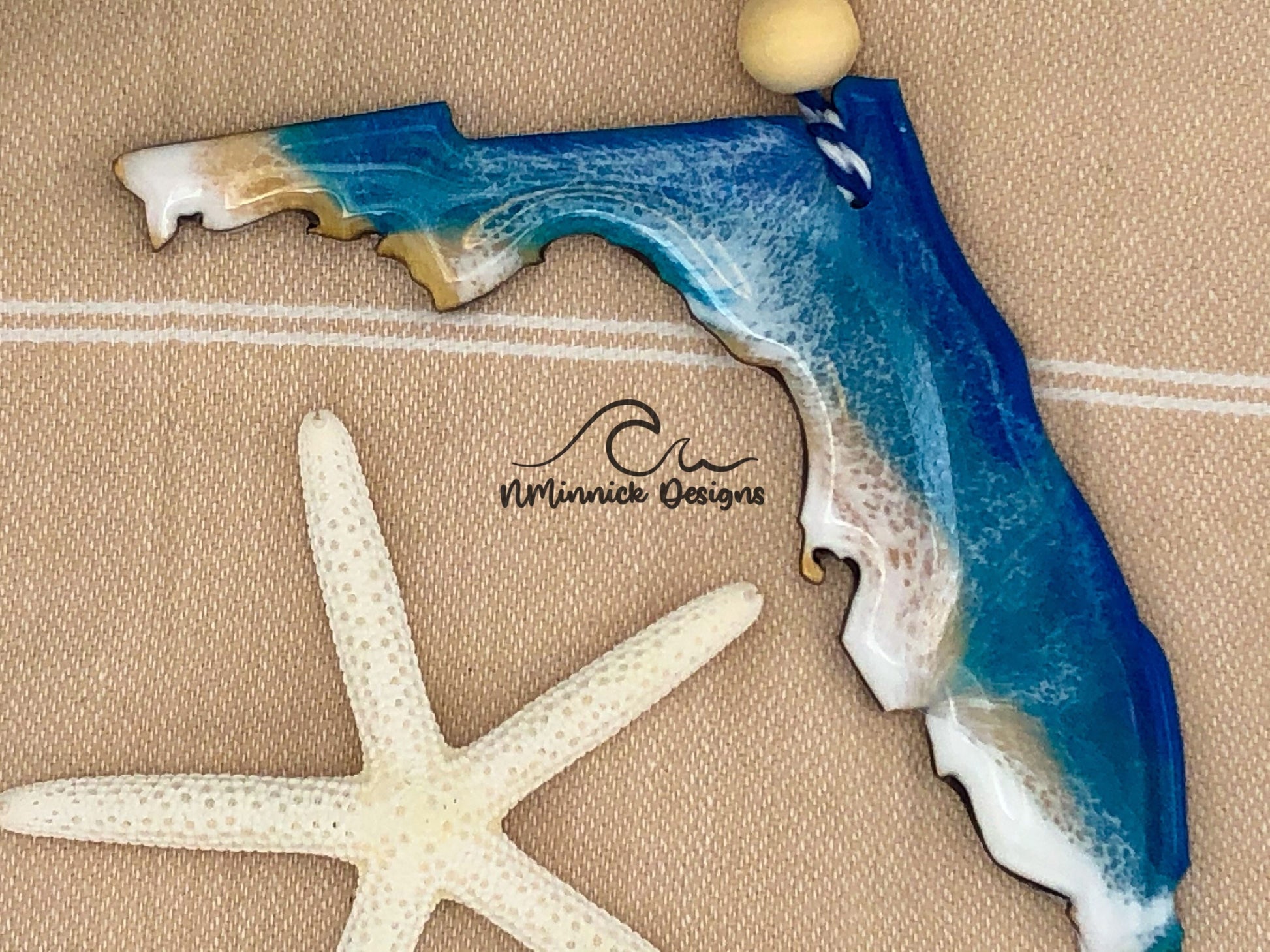 Florida-shaped wooden Christmas ornament covered in blue and teal colored ocean resin art resembling the waves along Florida's Atlantic Coast. Finished with blue and white baker's twine and two wooden beads.