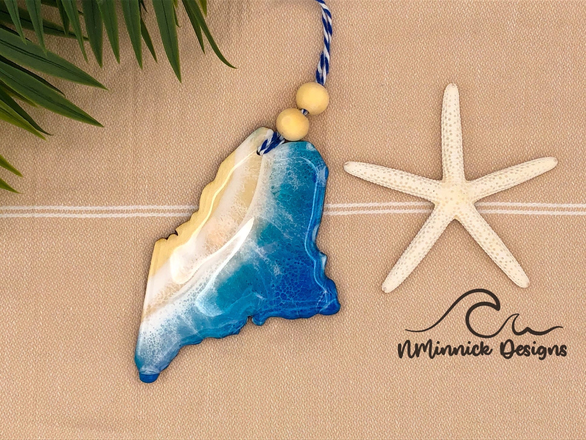 Maine-shaped wooden Christmas ornament covered in blue and teal colored ocean resin art resembling the waves along Maine's Atlantic Coast. Finished with blue and white baker's twine and two wooden beads.
