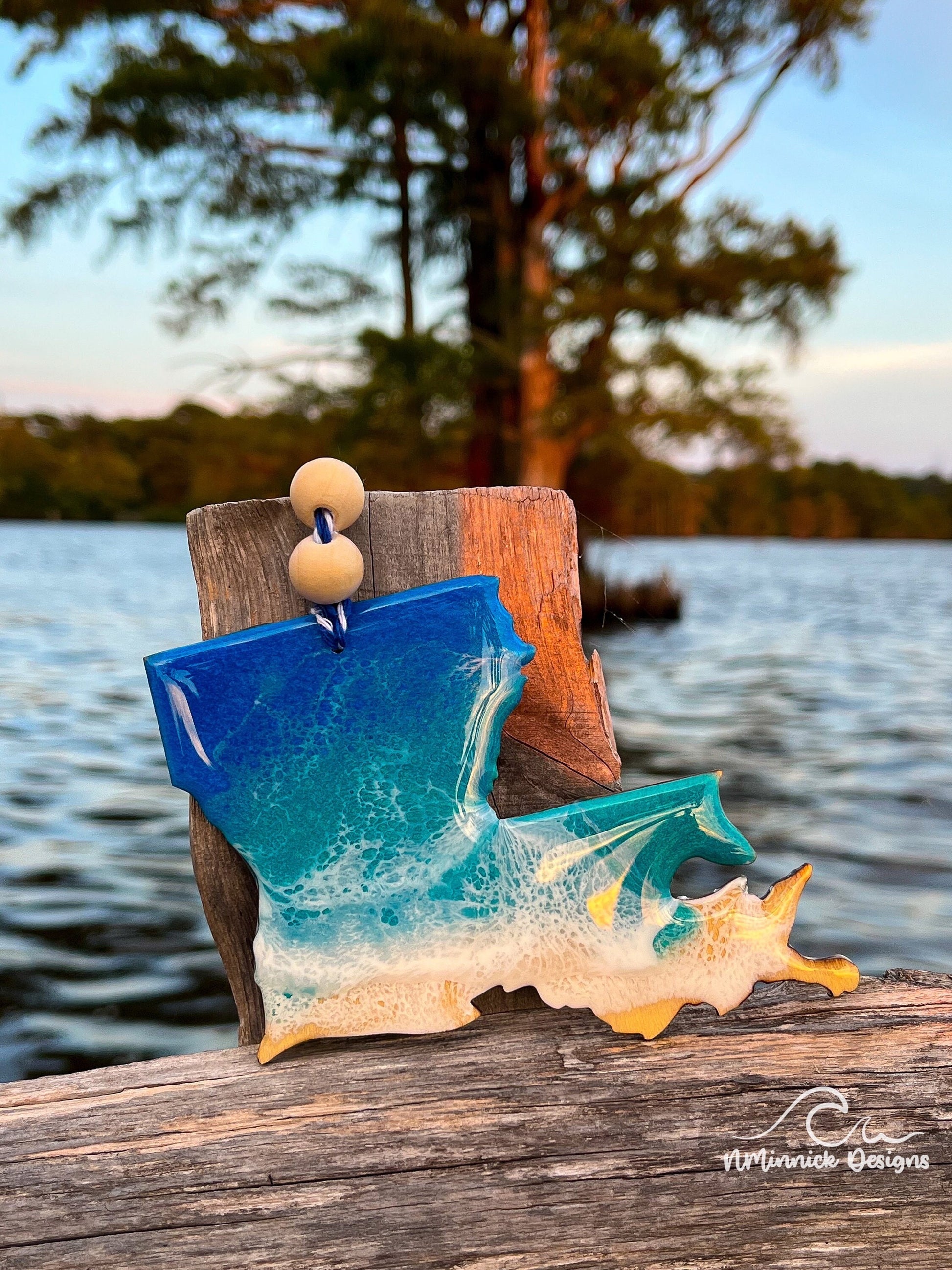 Louisiana-shaped wooden Christmas ornament covered in blue and teal colored ocean resin art resembling the waves along Louisiana's Gulf Coast. Finished with blue and white baker's twine and two wooden beads.