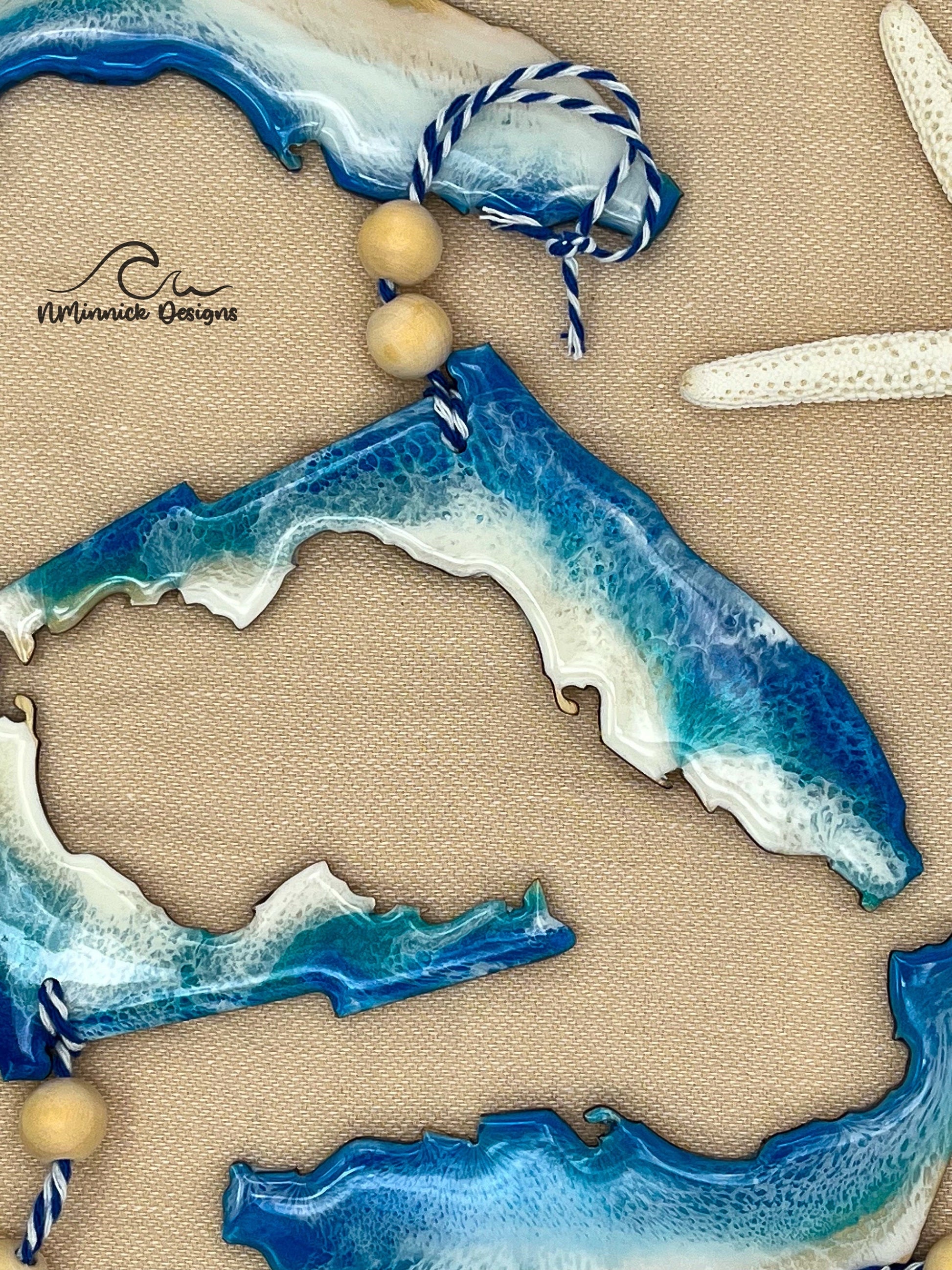 Florida-shaped wooden Christmas ornament covered in blue and teal colored ocean resin art resembling the waves along Florida&#39;s Gulf Coast. Finished with blue and white bakers twine and two wooden beads.