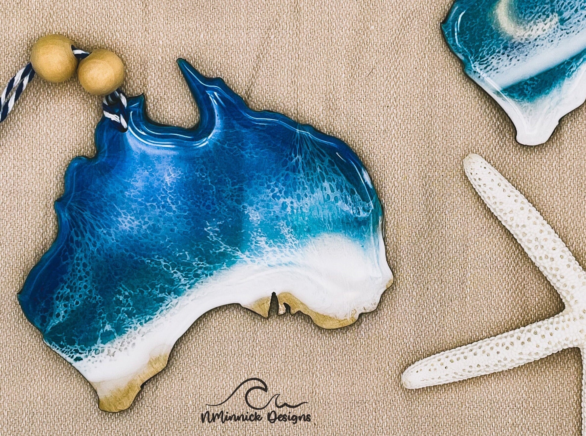 Australia-shaped wooden Christmas ornament covered in blue and teal ocean resin art resembling the waves of the Sunshine Coast. Finished with blue and white baker's twine and two wooden beads.