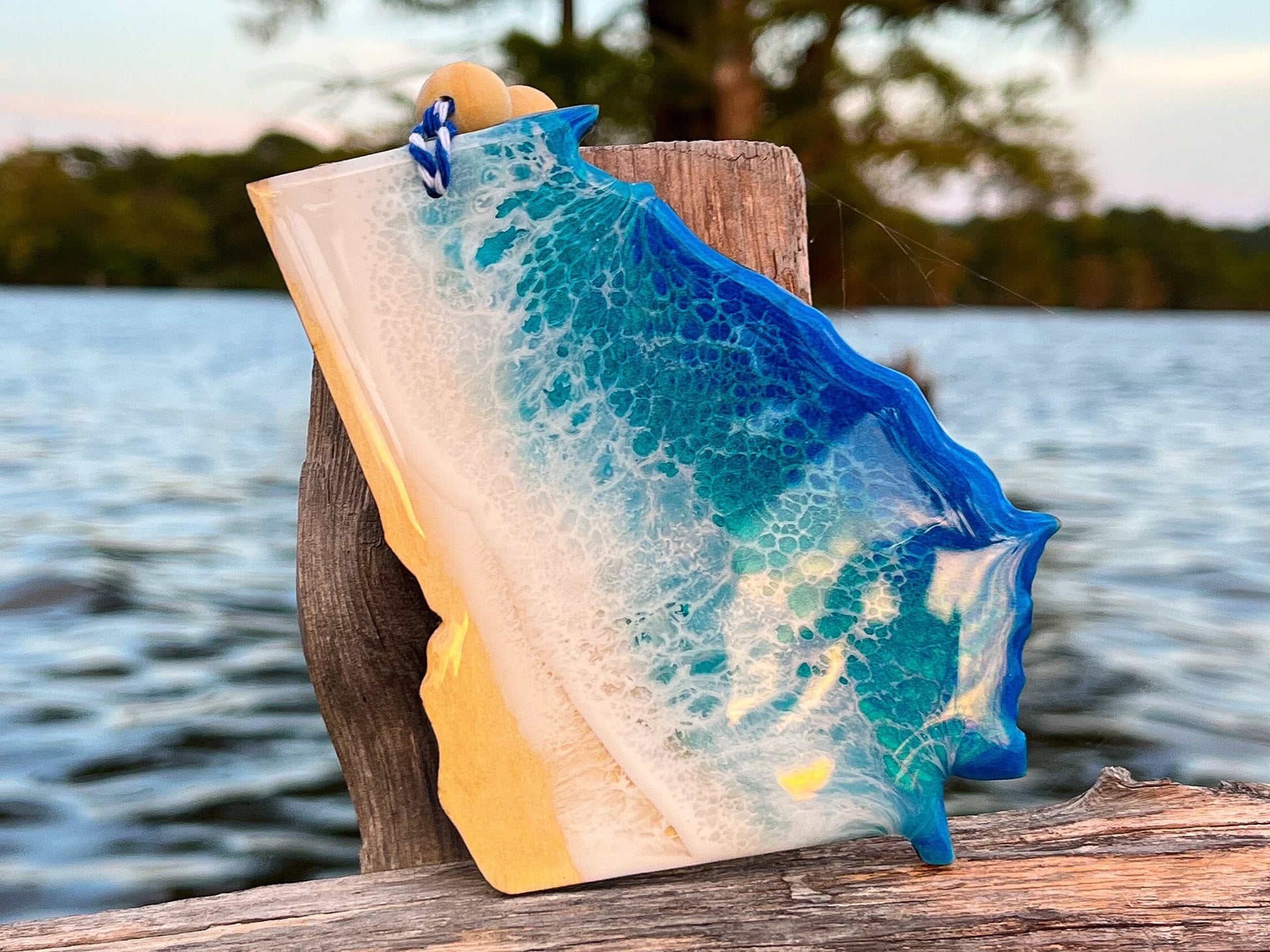 Georgia-shaped wooden Christmas ornament covered in blue and teal colored ocean resin art resembling the waves along Georgia's Coast. Finished with blue and white baker's twine and two wooden beads.