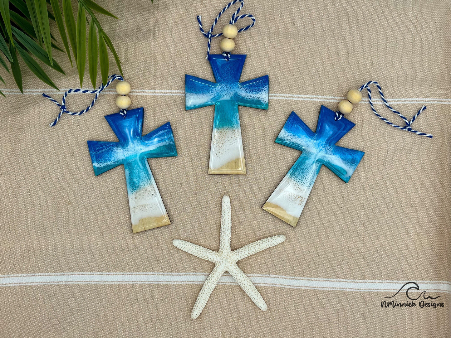 Three cross shaped wooden Christmas ornaments covered in blue ocean resin art, resembling ocean waves.  Finished with baker's twine and two wooden beads.  Laying on a tan beach towel next to a natural starfish and palm leaves.