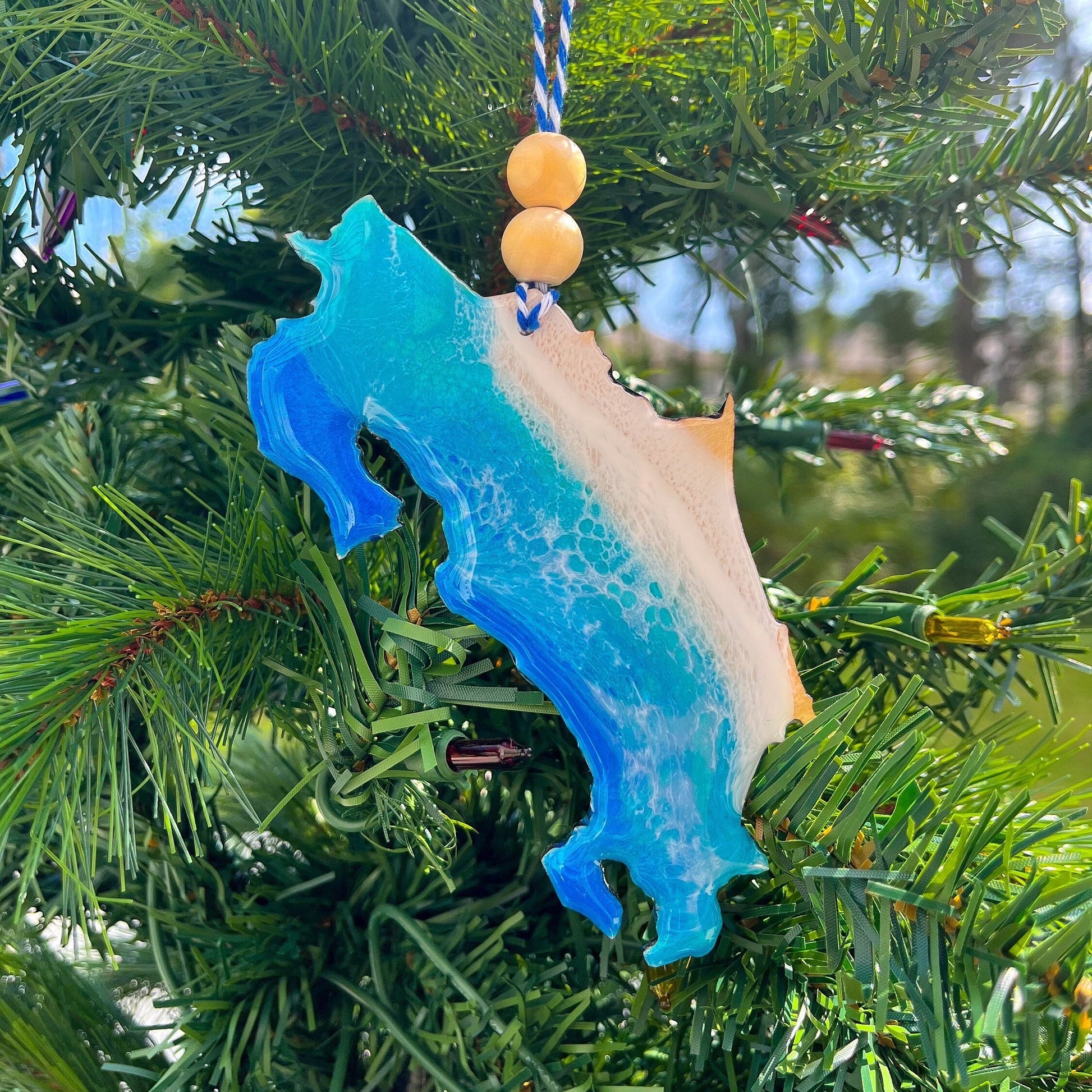 A Costa Rica shaped wooden Christmas ornament covered in blue ocean resin art, resembling the waves of the ocean.  Finished with bakers twine and two wooden beads and hanging on a Christmas tree.