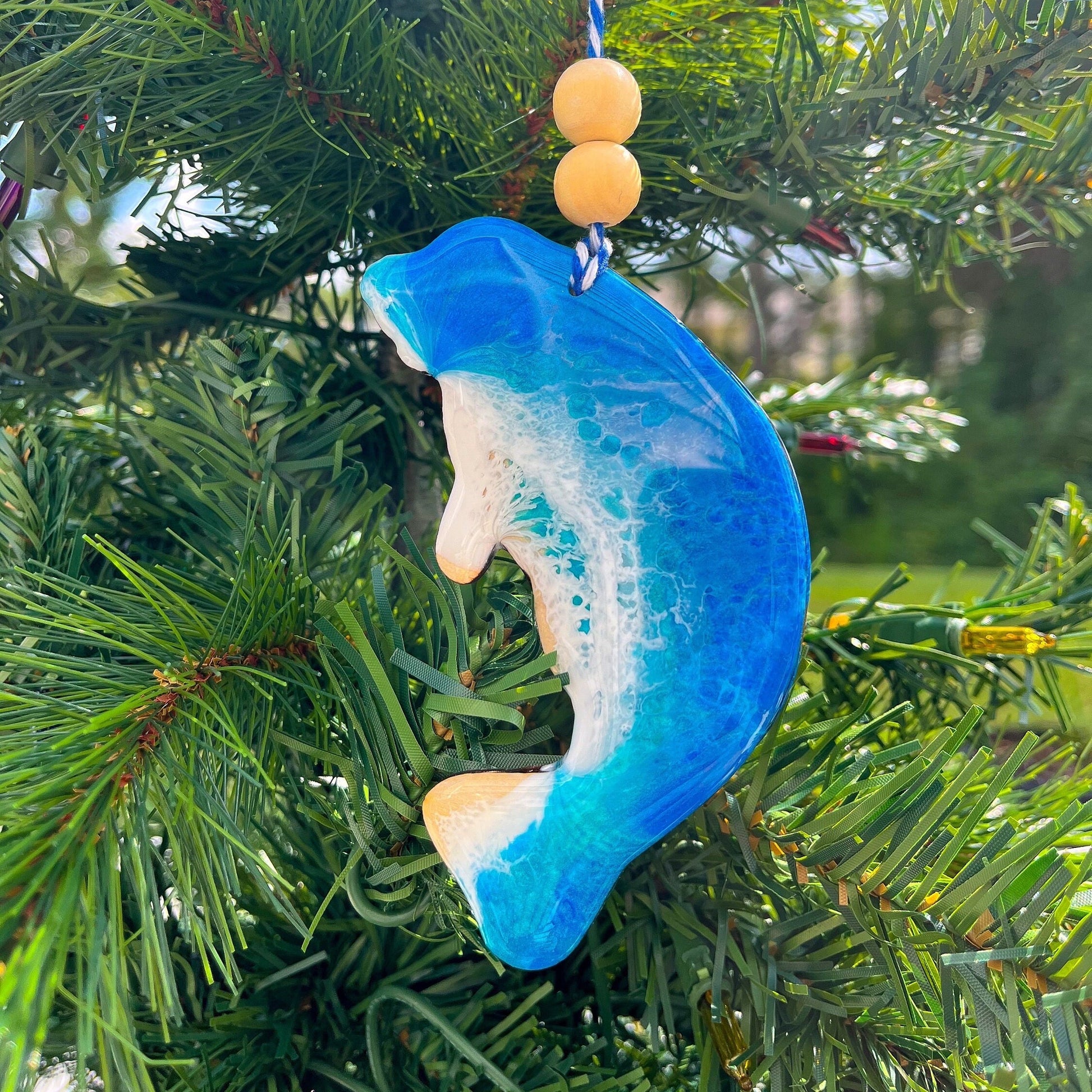 A manatee shaped wooden Christmas ornament covered in blue ocean resin art, resembling the waves of the ocean.  Finished with baker&#39;s twine and two wooden beads and hanging on a Christmas tree.