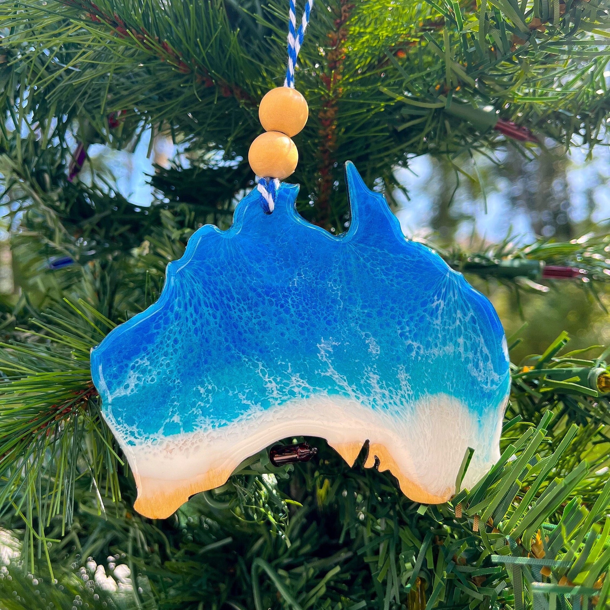 An Australia shaped wooden Christmas ornament covered in blue ocean resin art, resembling the waves of the ocean.  Finished with baker's twine and two wooden beads and hanging on a Christmas tree.