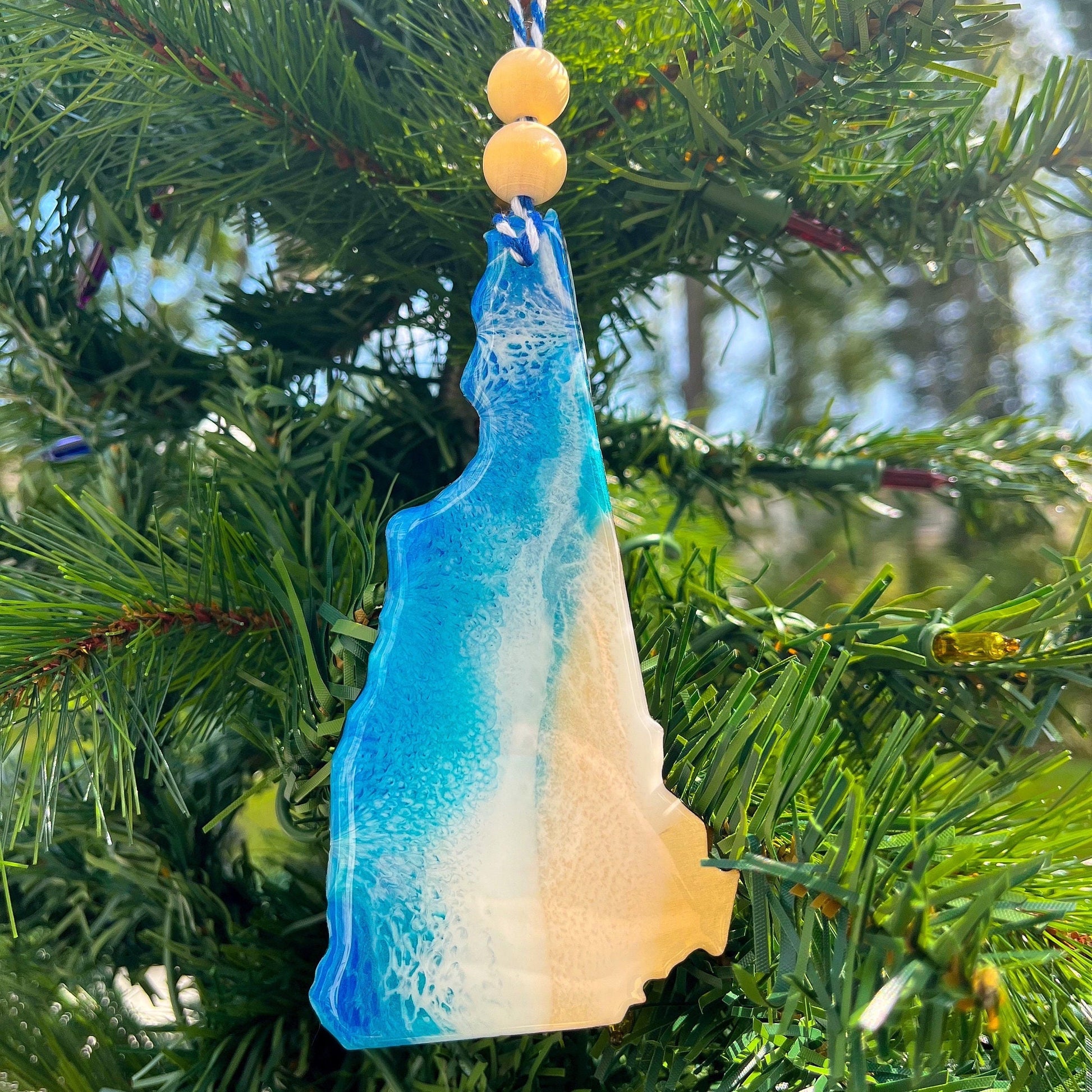 a new hampshire state shaped wooden Christmas ornament covered in ocean blue and okinawa blue ocean resin art resembling ocean waves against the shore.  Finished with baker's twine and two wooden beads and hanging on a christmas tree.