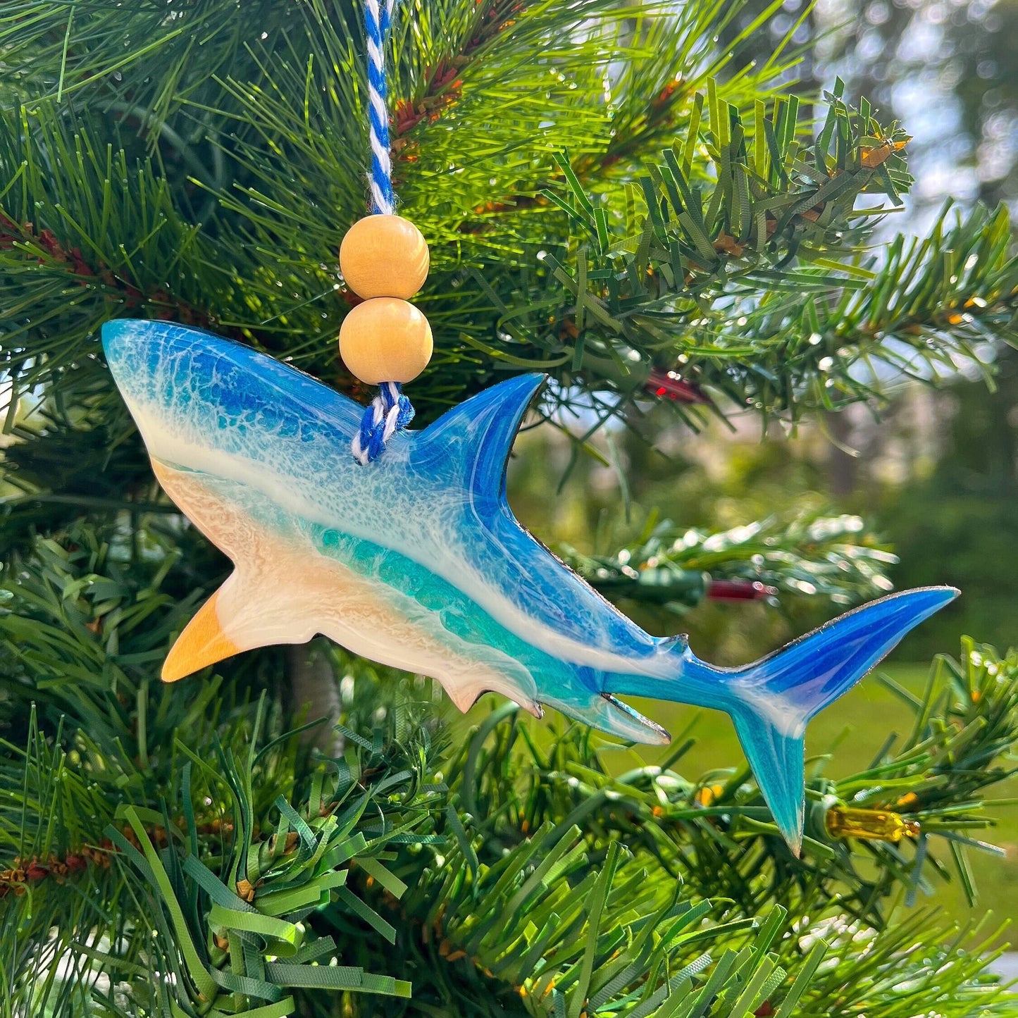 A shark shaped wooden Christmas ornament covered in blue ocean resin art, resembling the waves of the ocean. Finished with baker&#39;s twine and two wooden beads and hanging on a Christmas tree.