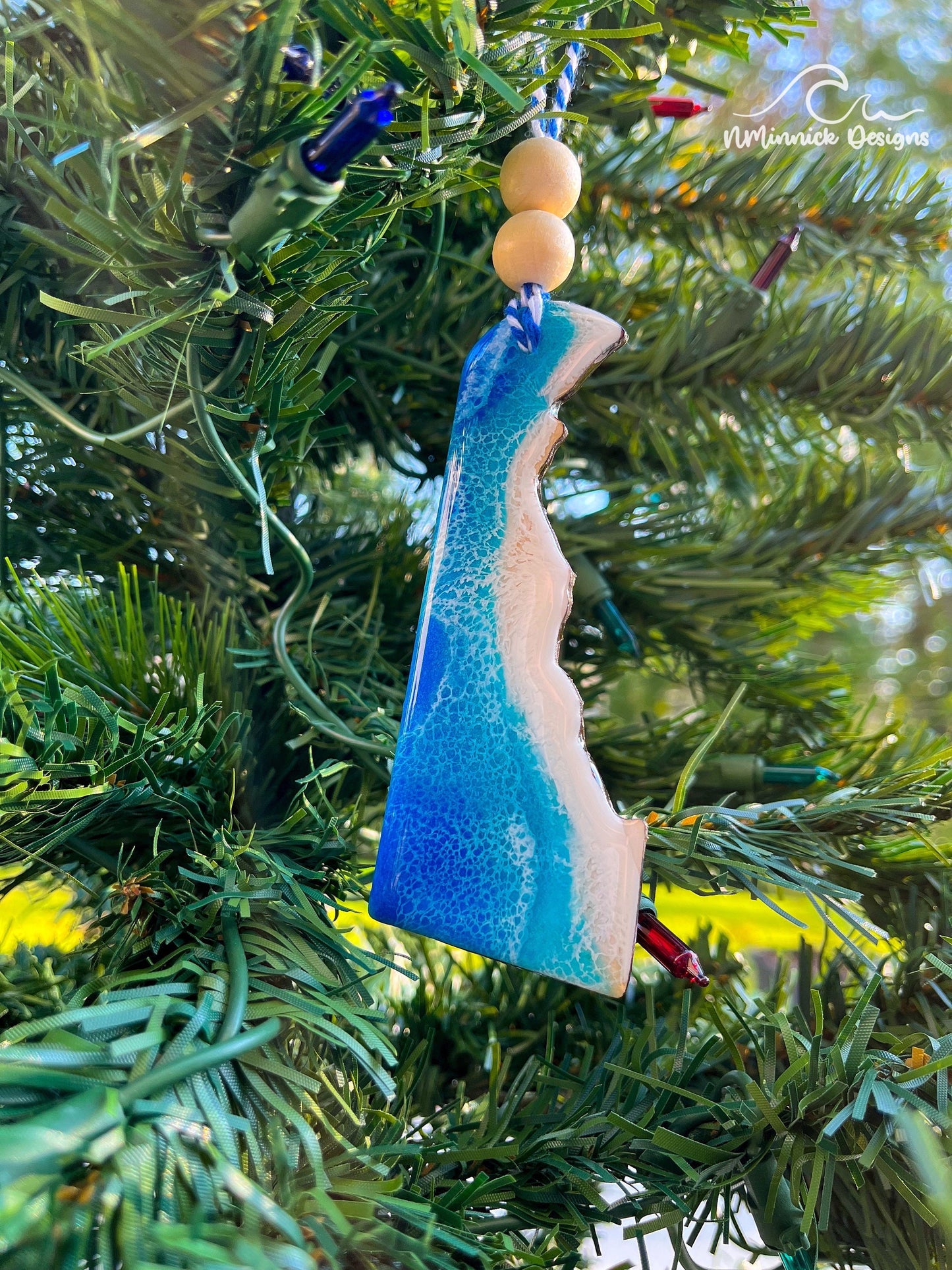 A blue and white resin ornament shaped like the state of Delaware, hanging on a tree with white and blue twine and wooden beads.