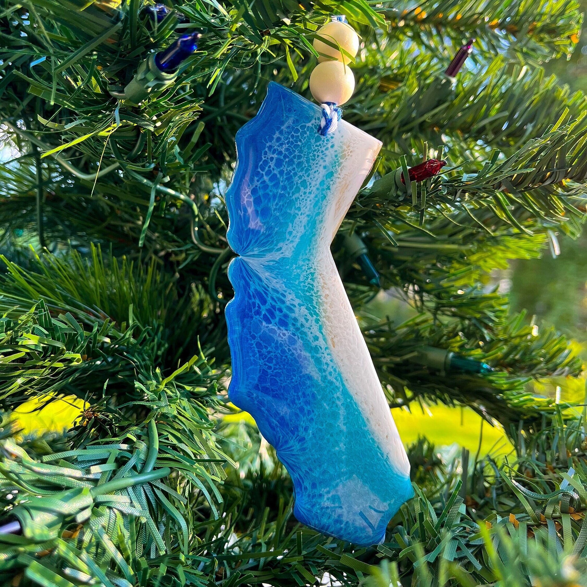 A california shaped wooden christmas ornament covered in layers of blue and teal ocean resin art resembling the waves of the ocean.  Finished with baker's twine and two wooden beads and hanging on a christmas tree.