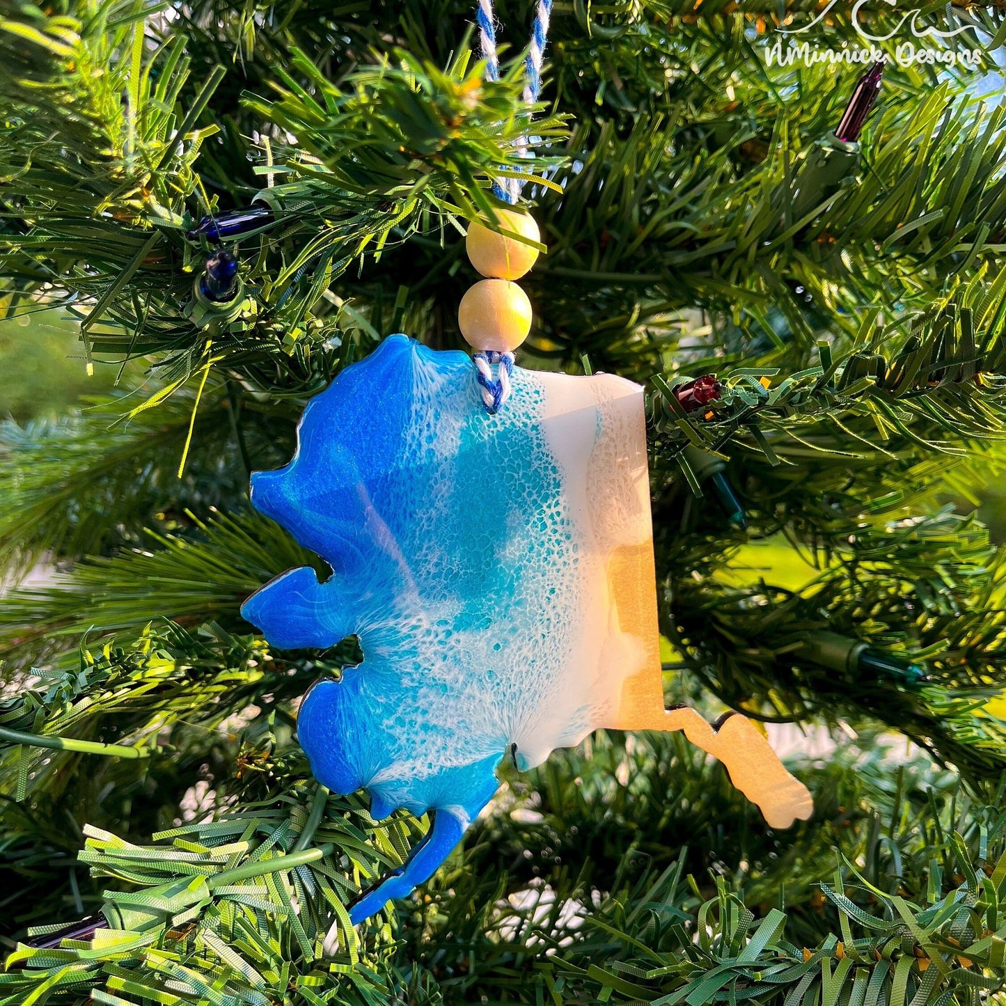 Alaska-shaped wooden Christmas ornament covered in blue and teal colored ocean resin art resembling the waves along Alaska's Pacific Coast. Finished with blue and white baker's twine and two wooden beads.
