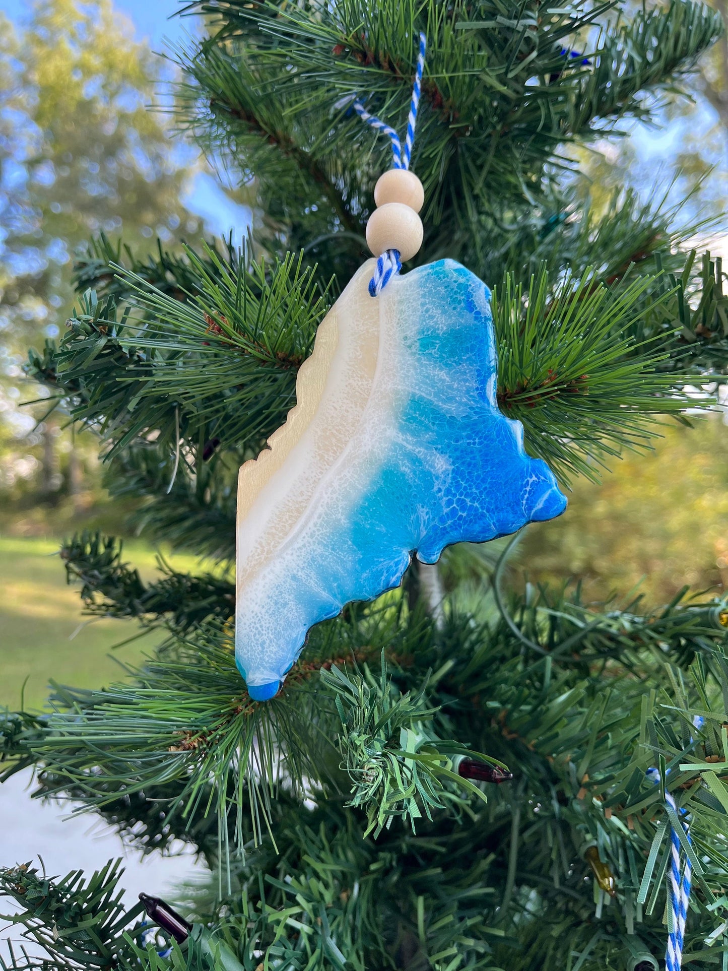 A blue and white ocean resin art ornament in the shape of Maine hanging on a tree
