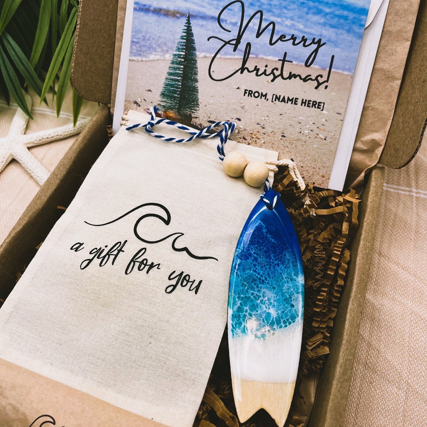 A Surfboard shaped wooden christmas ornament with ocean resin art, resembling the waves of the ocean. In a box with an ornament keepsake bag with a wave that says "a gift for you" and a Merry Christmas custom card.