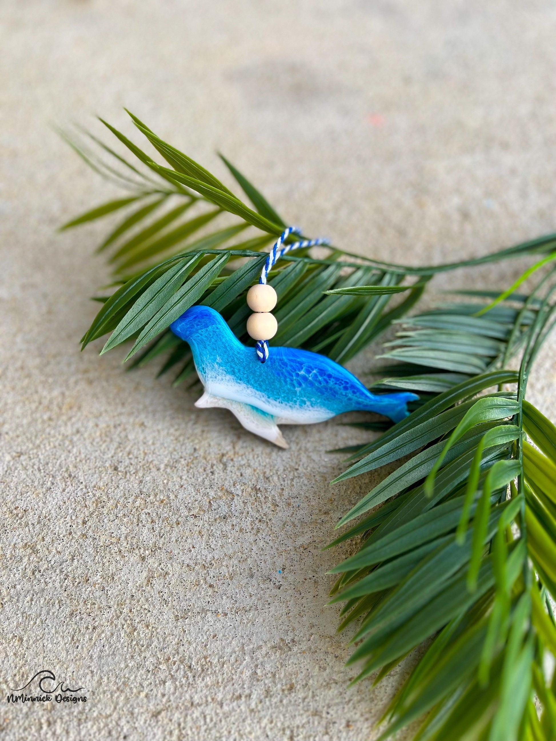 Harbor seal shaped wooden ornament with ocean resin waves on the front hanging from blue and white twine with two wooden beads and laying against palm leaves