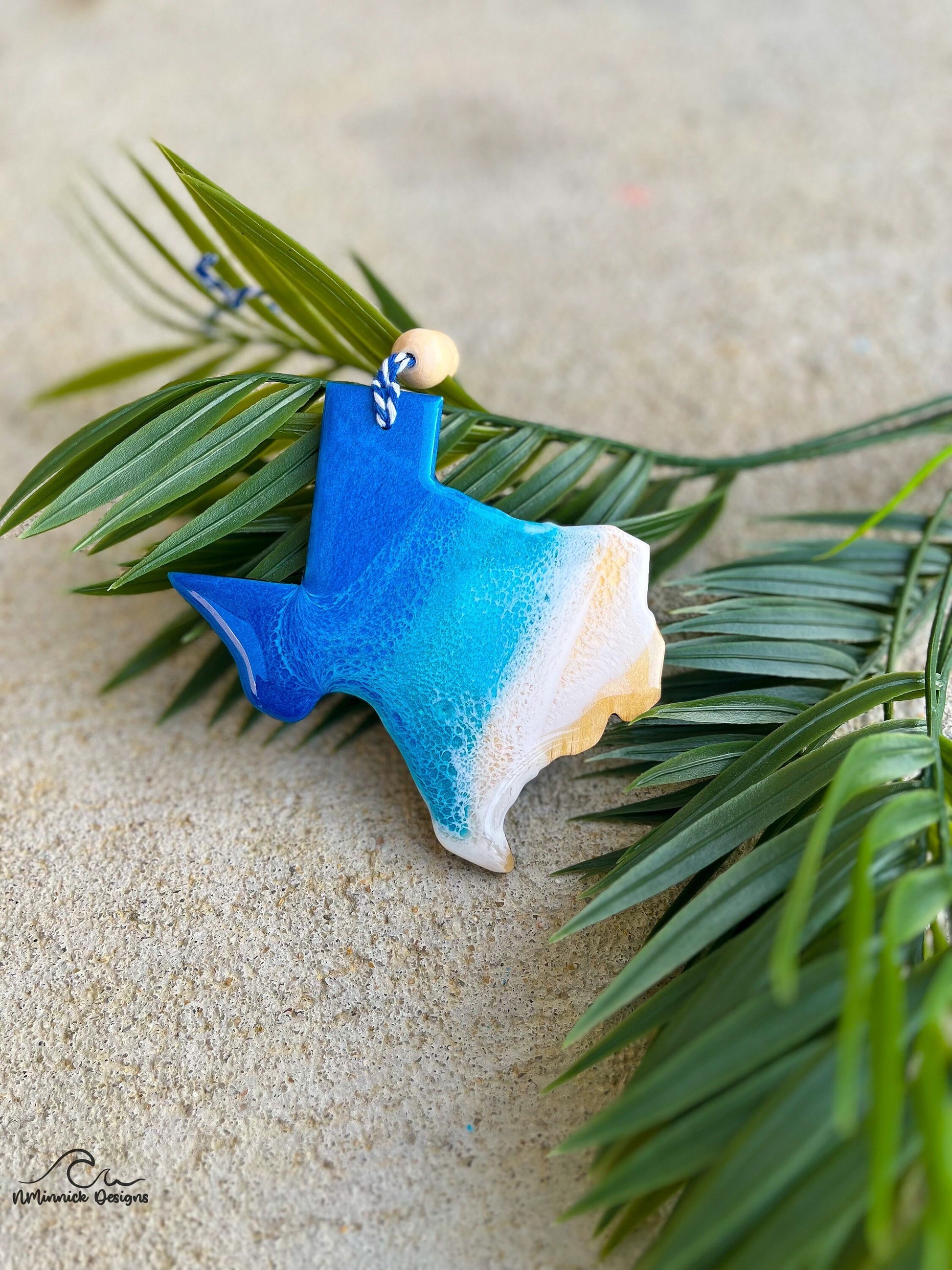 Texas shaped wooden ornament with ocean resin waves on the front hanging from blue and white twine with two wooden beads and laying against palm leaves