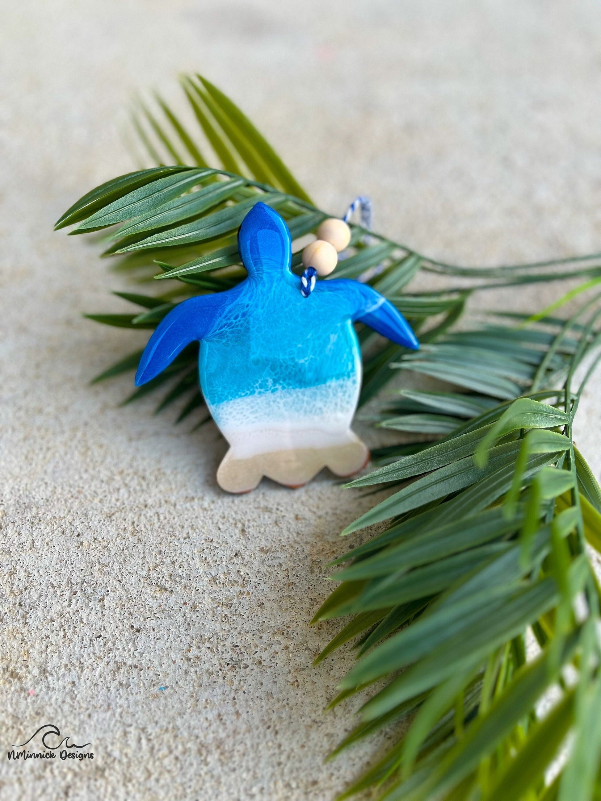 Sea Turtle shaped wooden ornament with ocean resin waves on the front hanging from blue and white twine with two wooden beads and laying against palm leaves