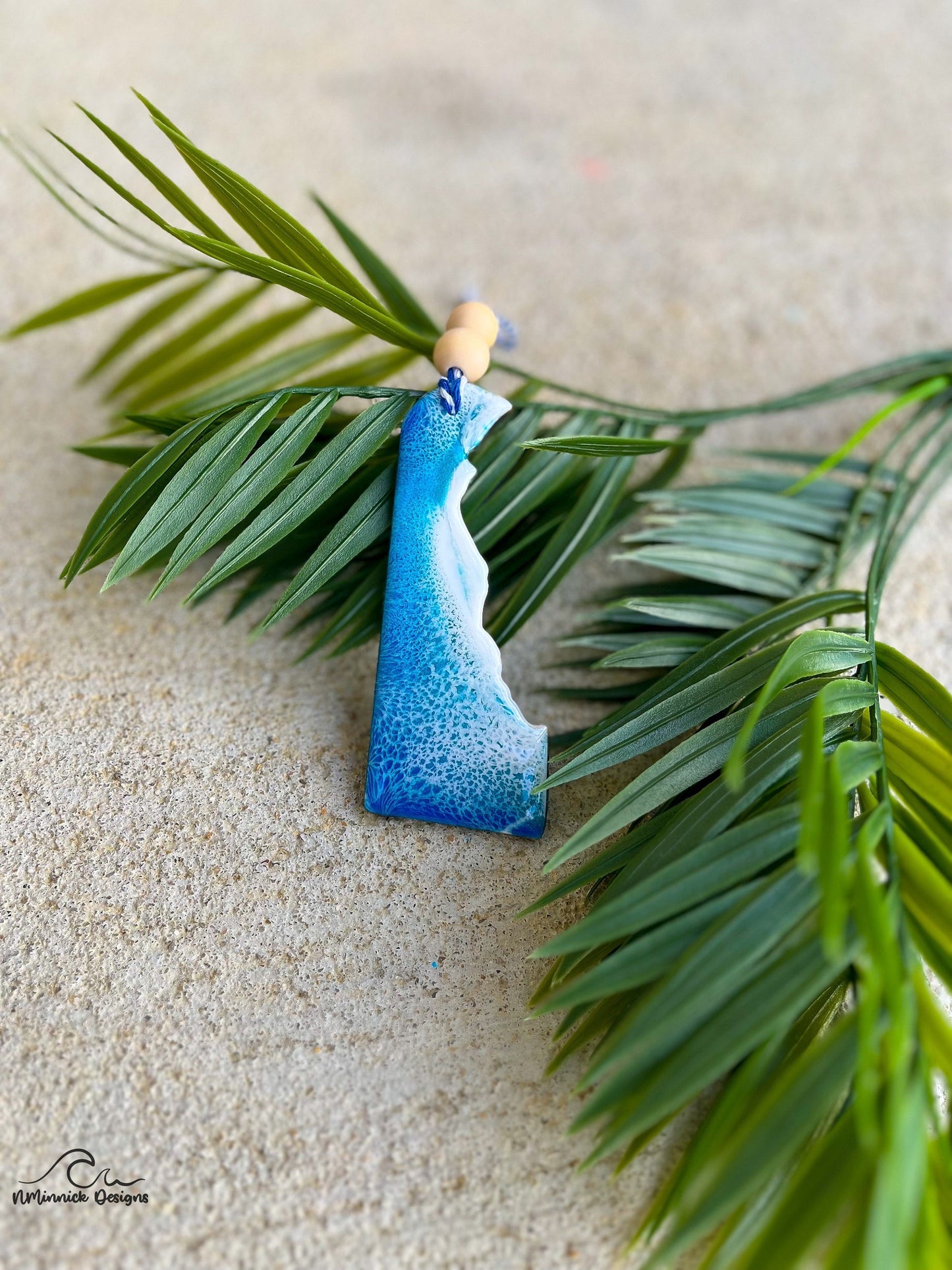 A delaware shaped wooden ornament with blue and white ocean resin art, finished with a strand of baker's twine and two wooden beads.