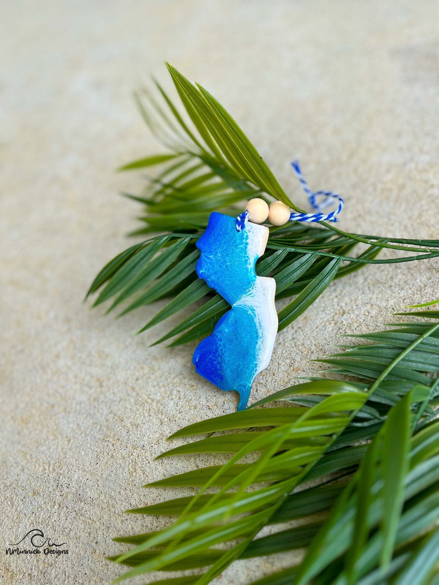 A New Jersey shaped wooden ornament with ocean resin waves on the front, laying against a palm leaf